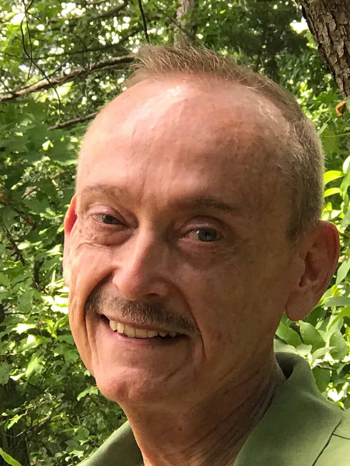 A smiling senior-aged man with short hair and a mustache, outdoors among green trees and leaves.