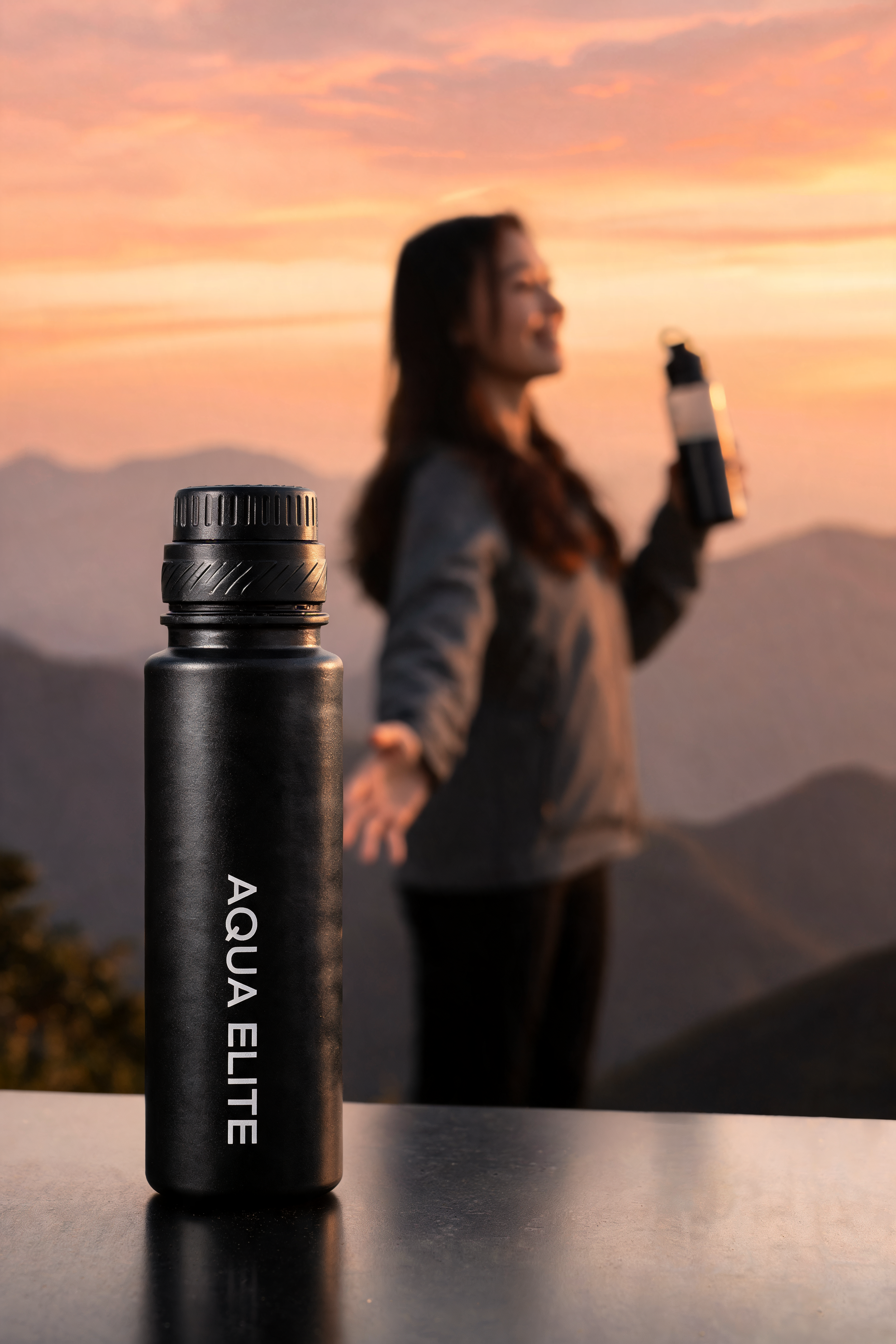 Close-up of a black water bottle labeled 'AQUA ELITE' on a smooth table surface. In the background, a woman with long hair is holding a water bottle, smiling, with a sunset sky and mountains behind her.