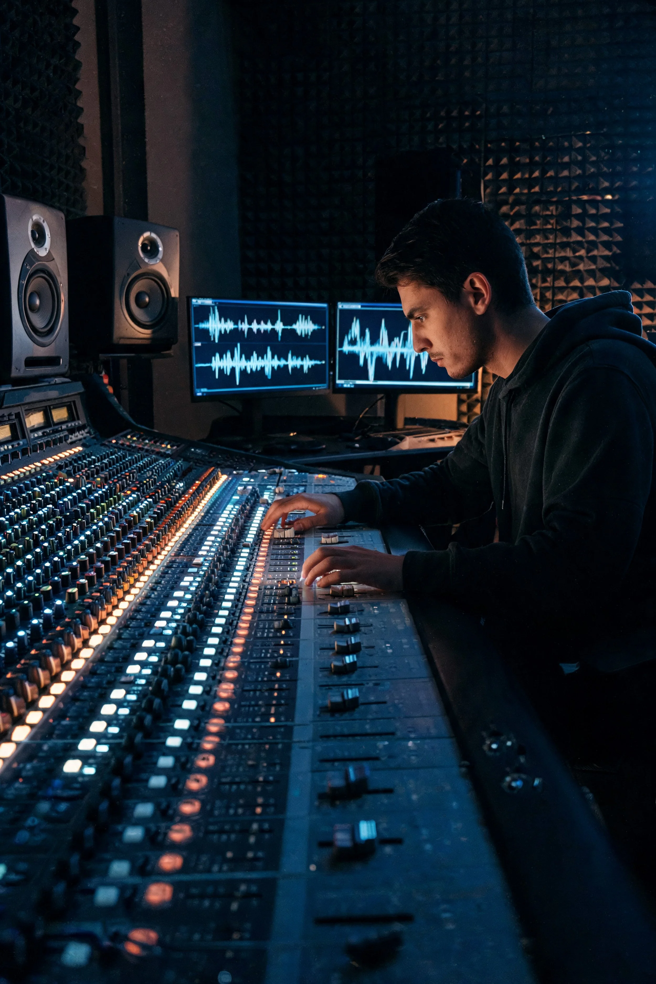 A man working in a music recording studio, working on a mixing console with two computer monitors displaying waveforms, and two studio speakers in the background.