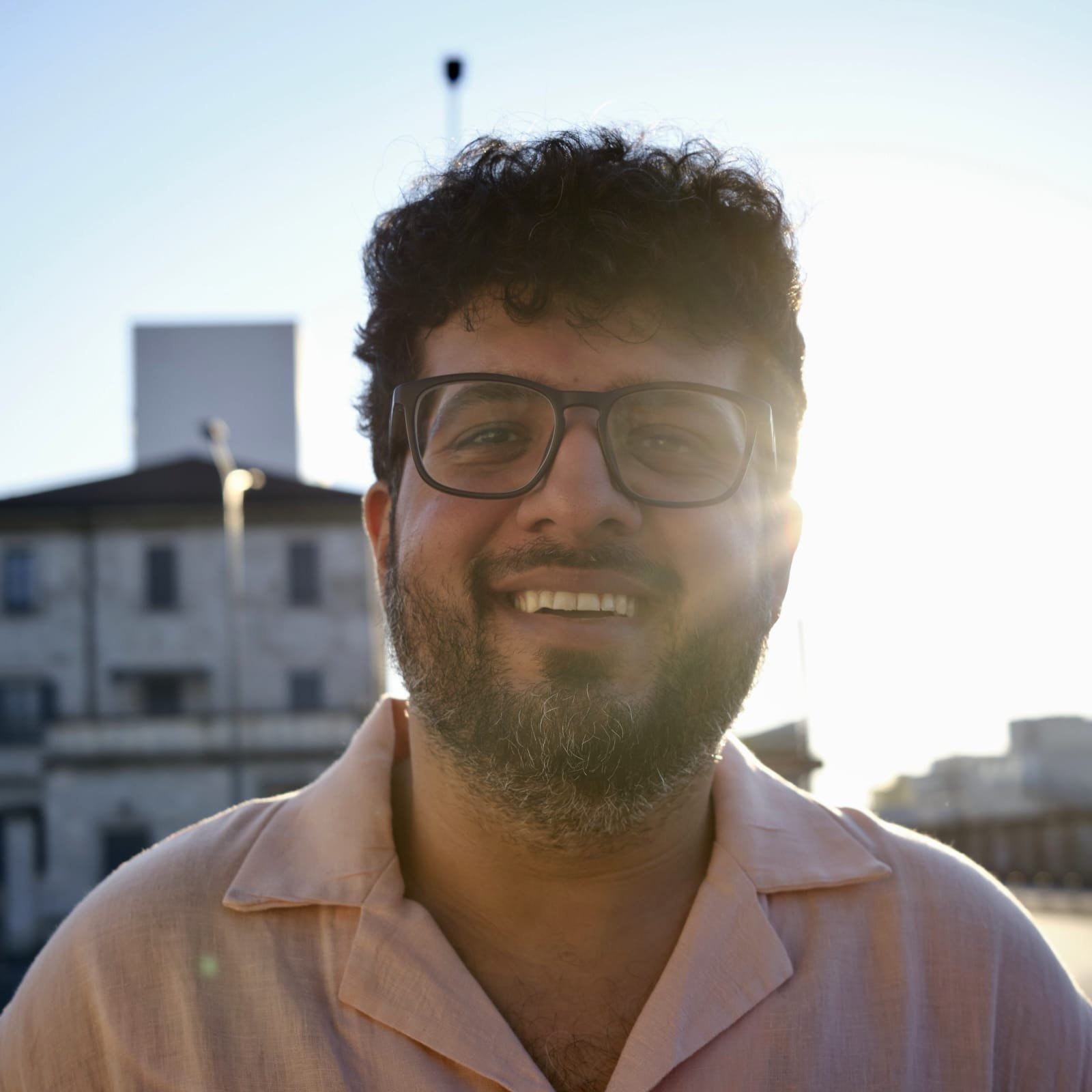 A smiling man with glasses smiling at the camera with sunlight in the background and buildings behind him.