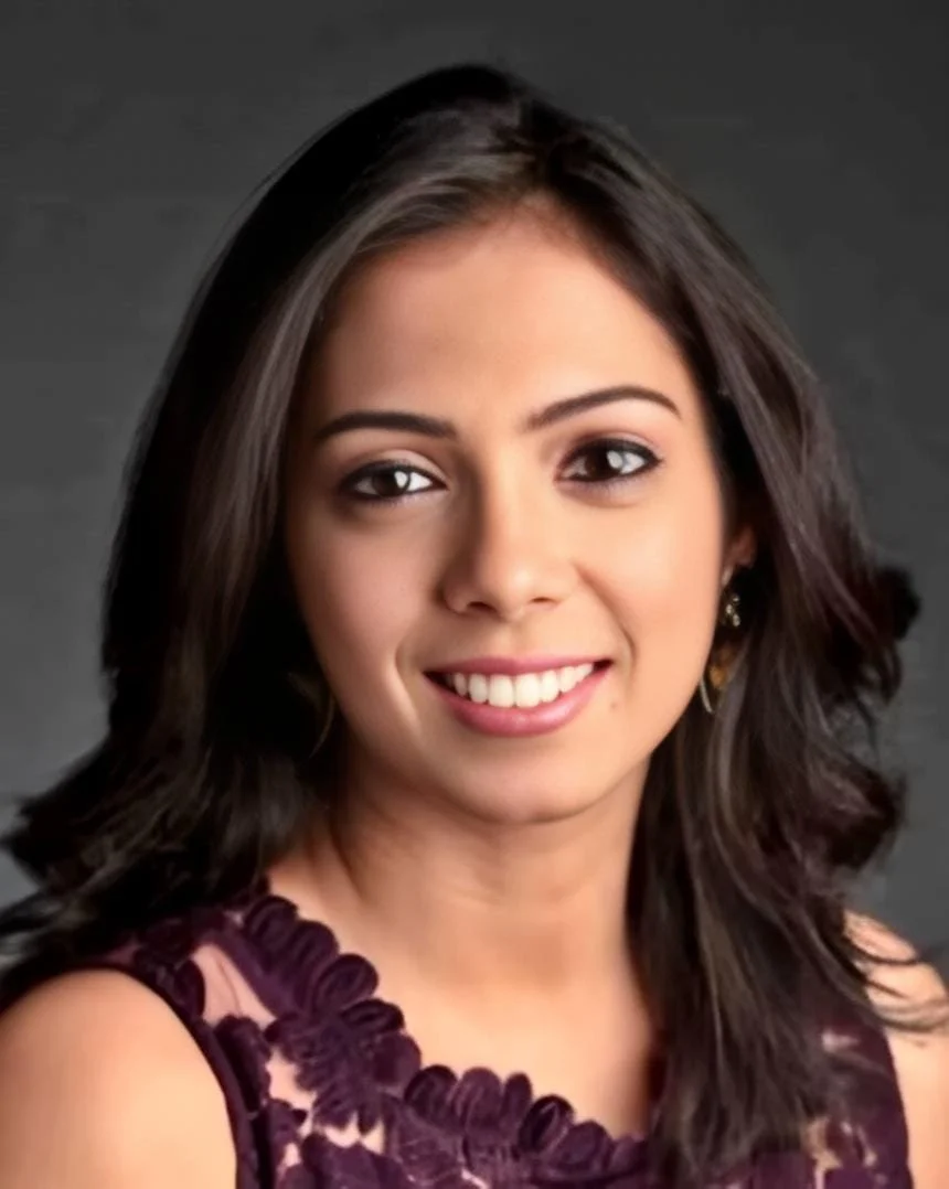 A young woman with shoulder-length dark hair, wearing a dark purple dress with lace details, smiling at the camera against a dark background.