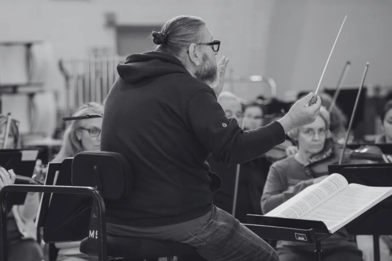 Conductor Daniel Henriks leading musicians during a rehearsal, with sheet music on stands and musicians in the background.