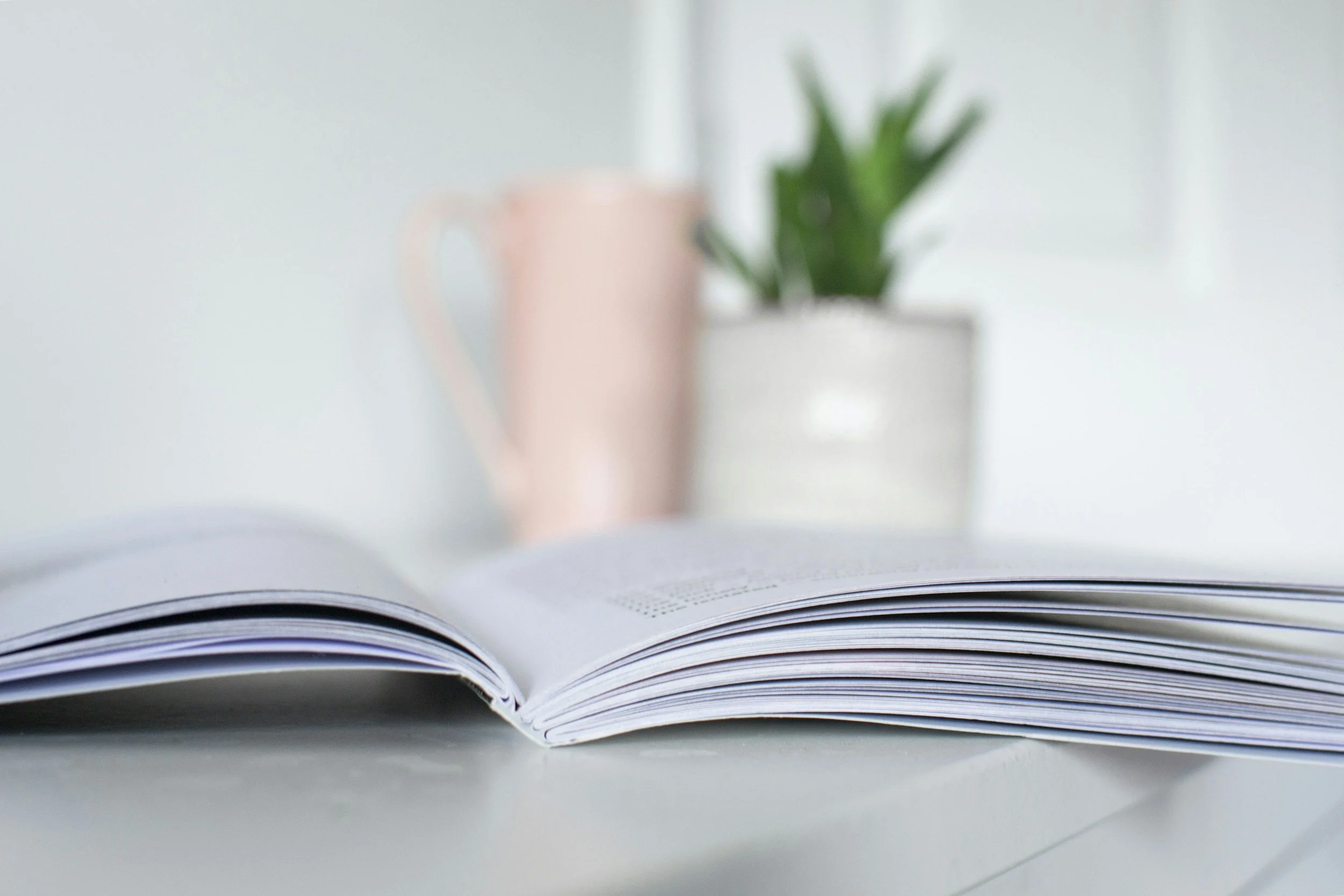 Open magazine on a table with a blurred background of a potted plant and a pink mug.