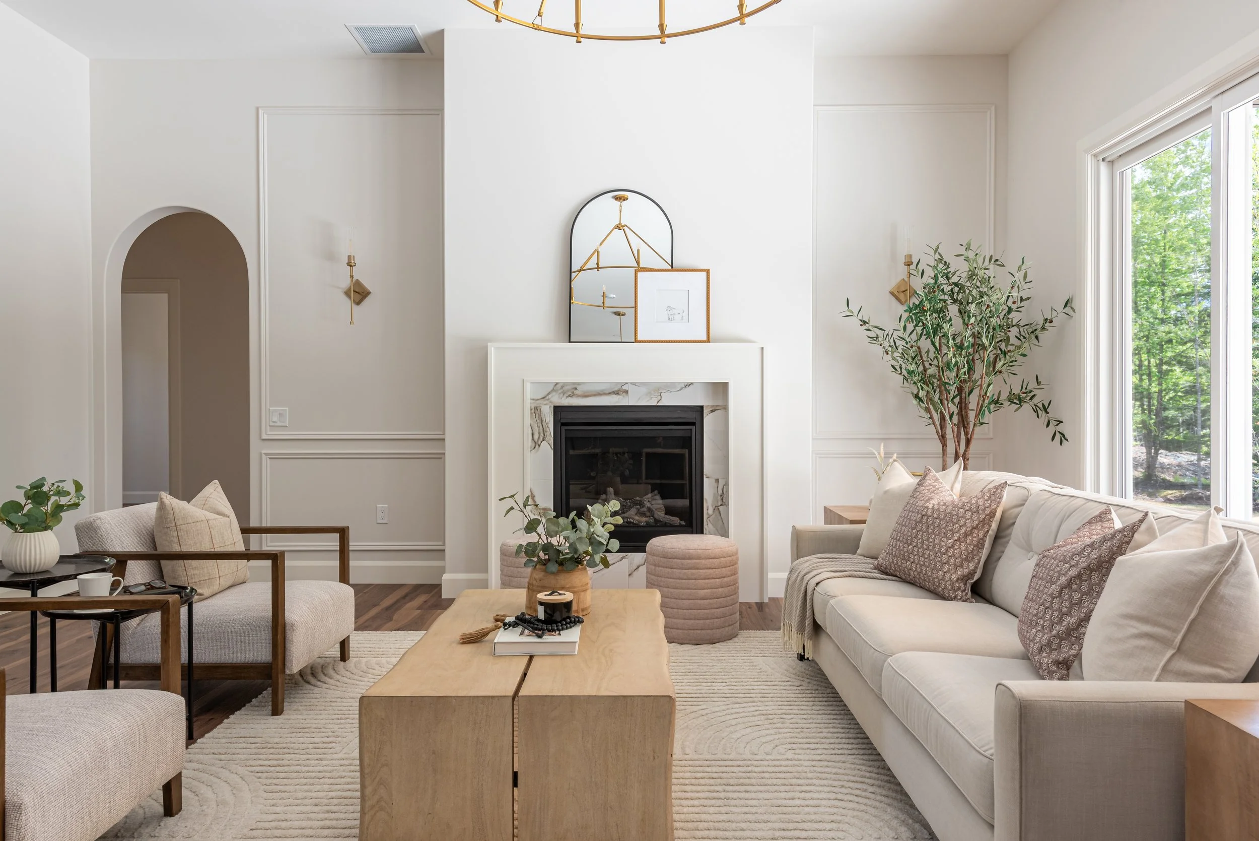 A living room with white walls, a fireplace with marble surround, a large window showing green trees, a beige sofa with patterned pillows, a coffee table with decor items, a side table with a plant, and modern wall sconces.