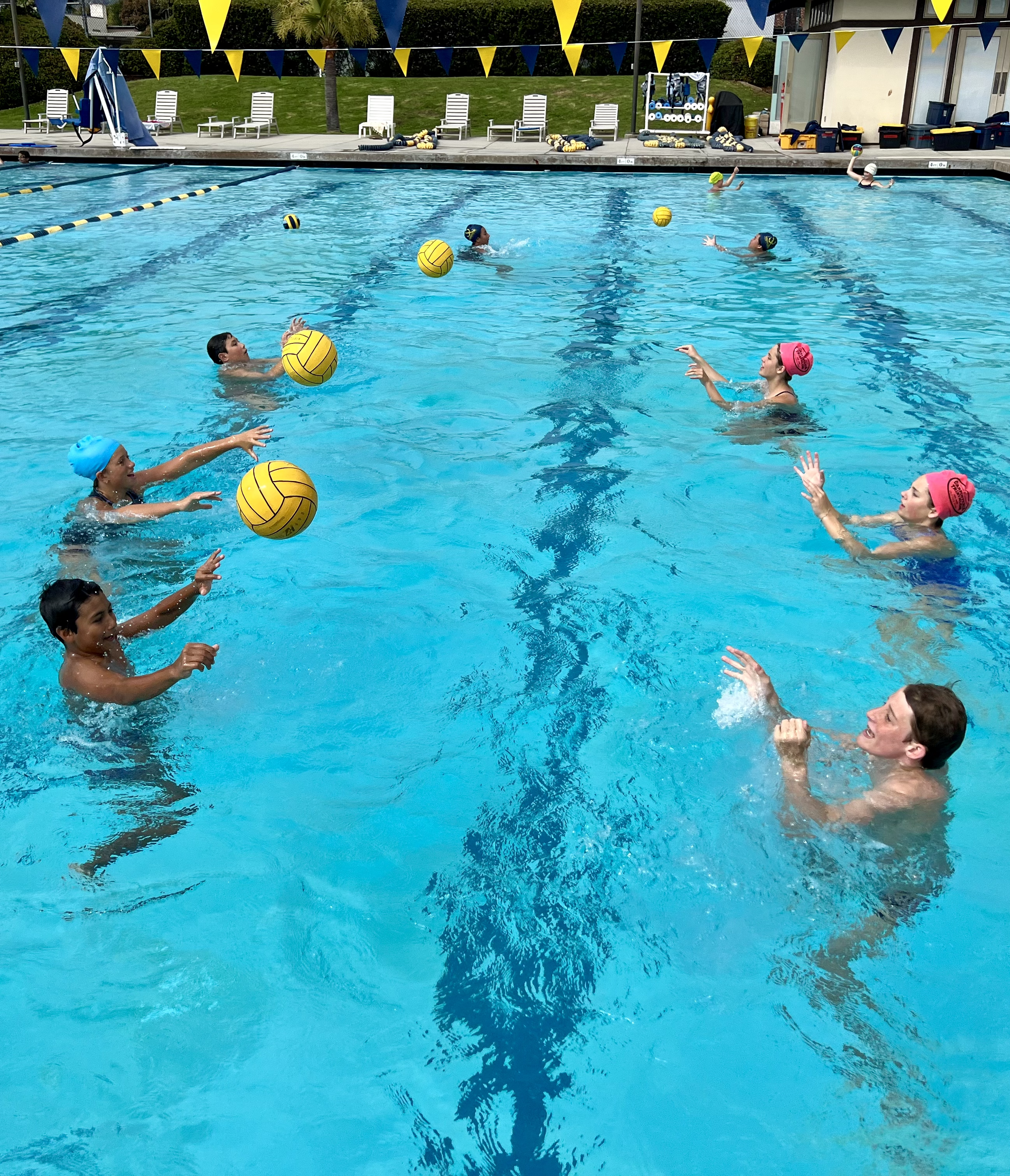 Children playing water polo in an outdoor swimming pool, with yellow and blue flags hanging above, and poolside chairs, equipment, and storage in the background.