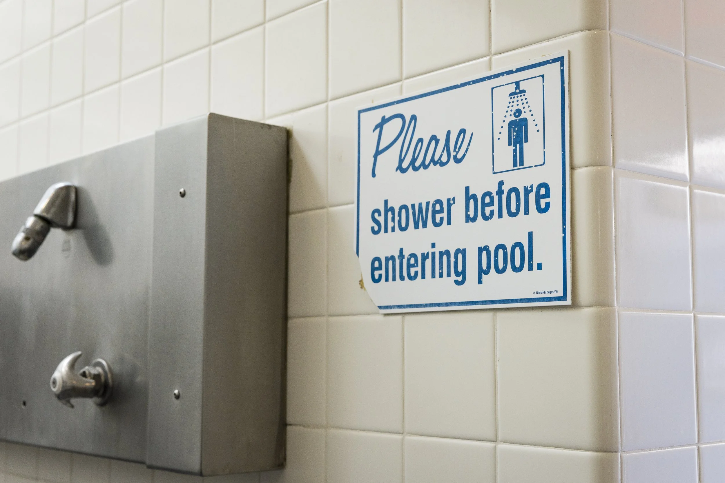 Broken sign on tiled bathroom wall instructing to shower before entering pool, with blue and white text and a shower icon.