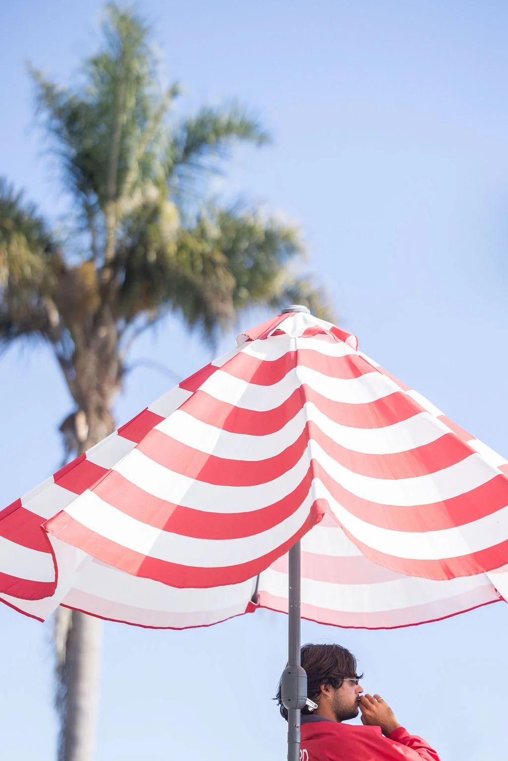 A pool lifeguard wearing sunglasses and a red jacket sitting under a red and white striped umbrella with a palm tree and clear blue sky in the background.