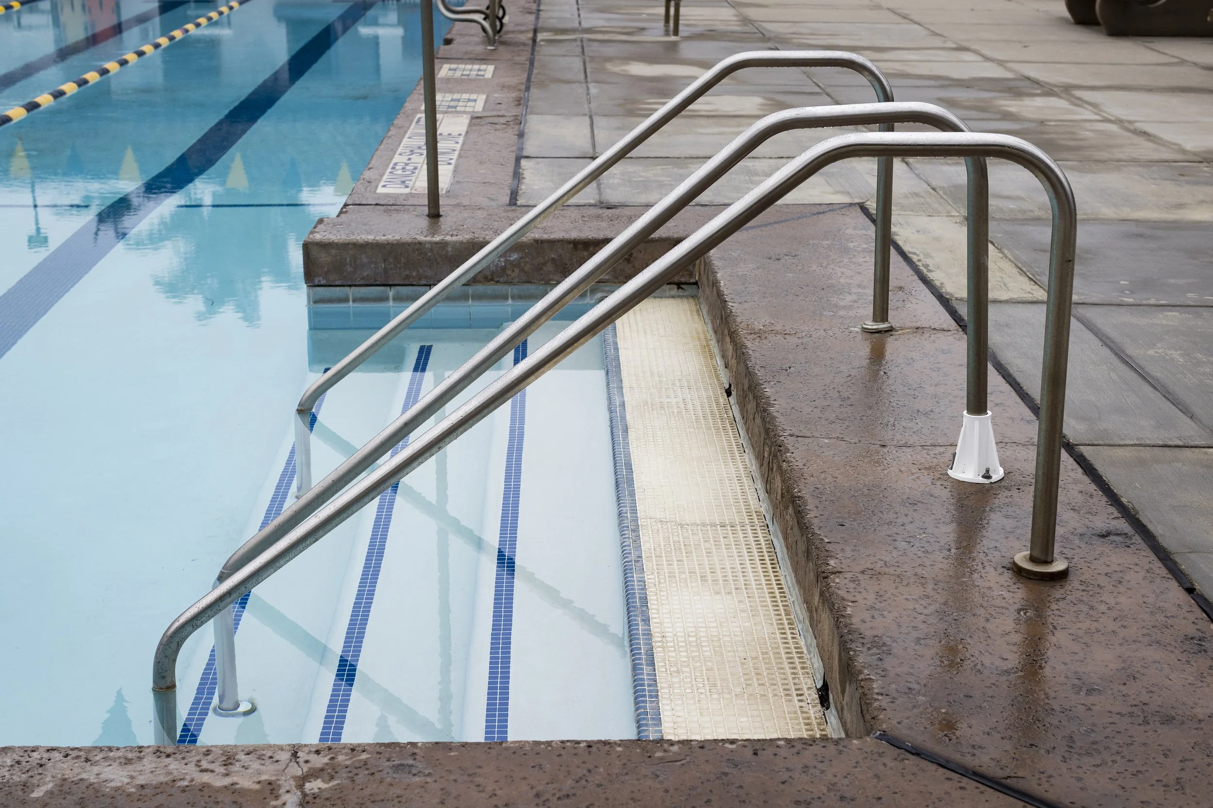 Empty outdoor swimming pool with metal handrails leading into the water and a tiled surrounding deck area.