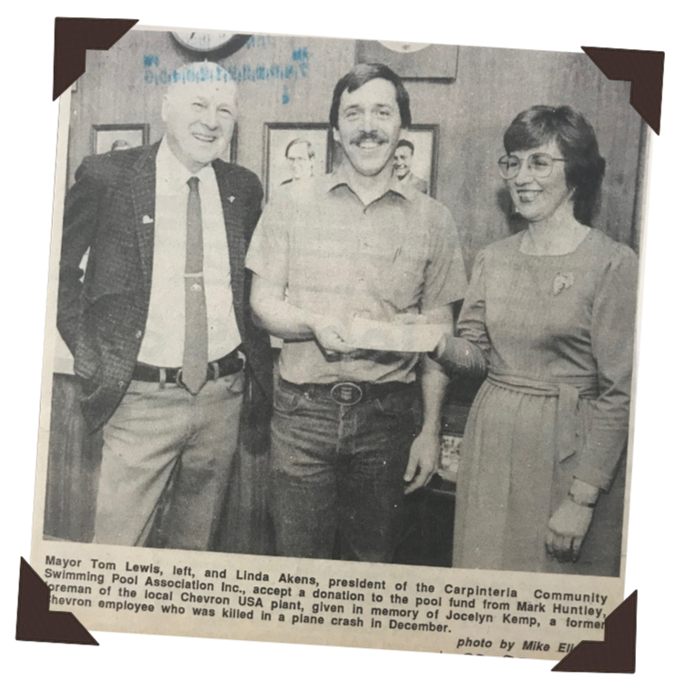 Black and white photo of three people indoors, with a framed picture behind. The man on the left is Mayor Tom Lewis, the man in the middle is Mark Huntley, and the woman on the right is Linda Akens. They are accepting a donation, and the woman is holding a check or document, smiling. The photo has a caption mentioning the donation to the Carpinteria Community Swimming Pool from Mark Huntley in memory of Jocelyn Kemp.