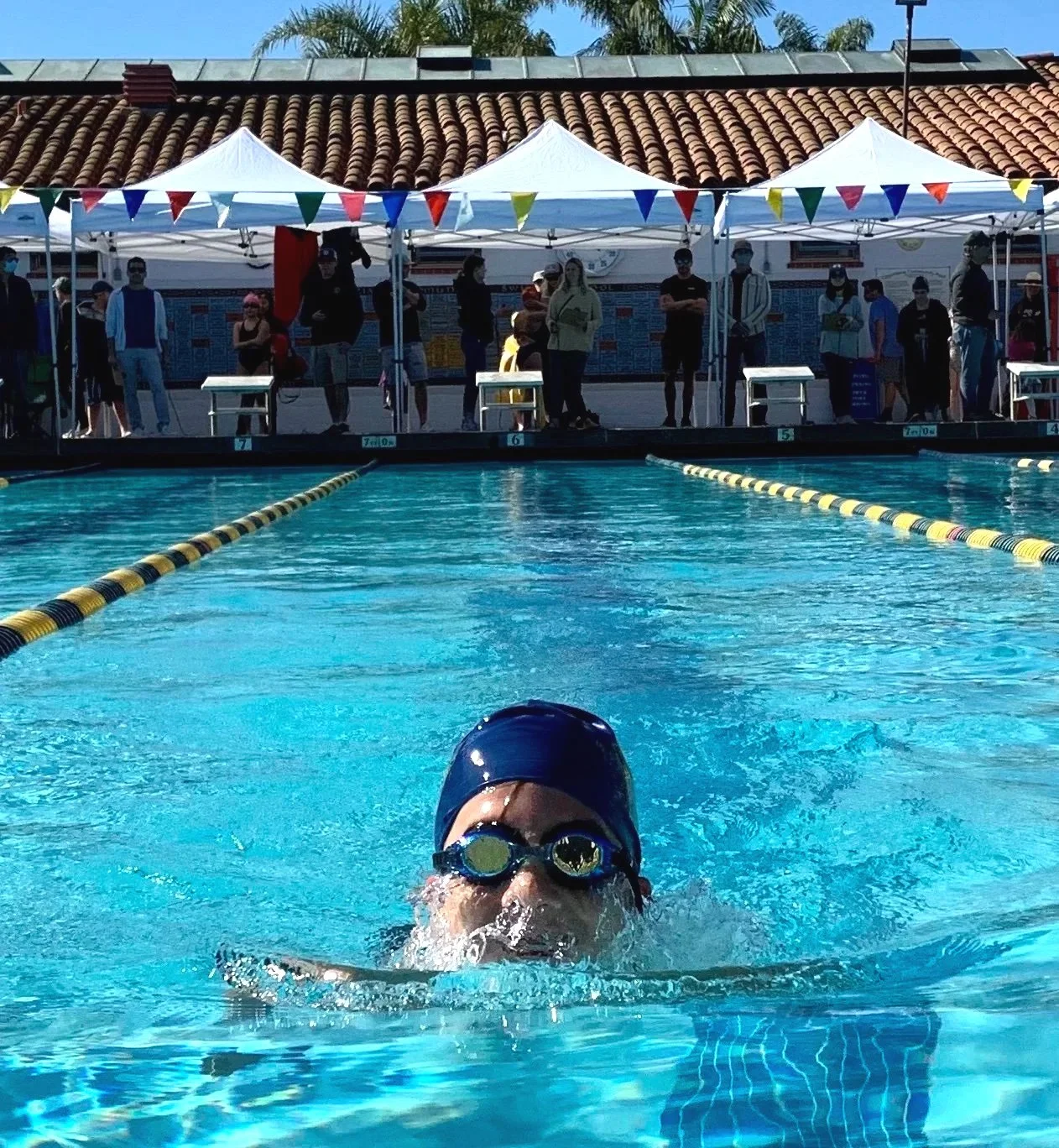 Swimmer in the water pool near the edge, wearing a swim cap and goggles, with a group of people and white tents with bunting in the background.