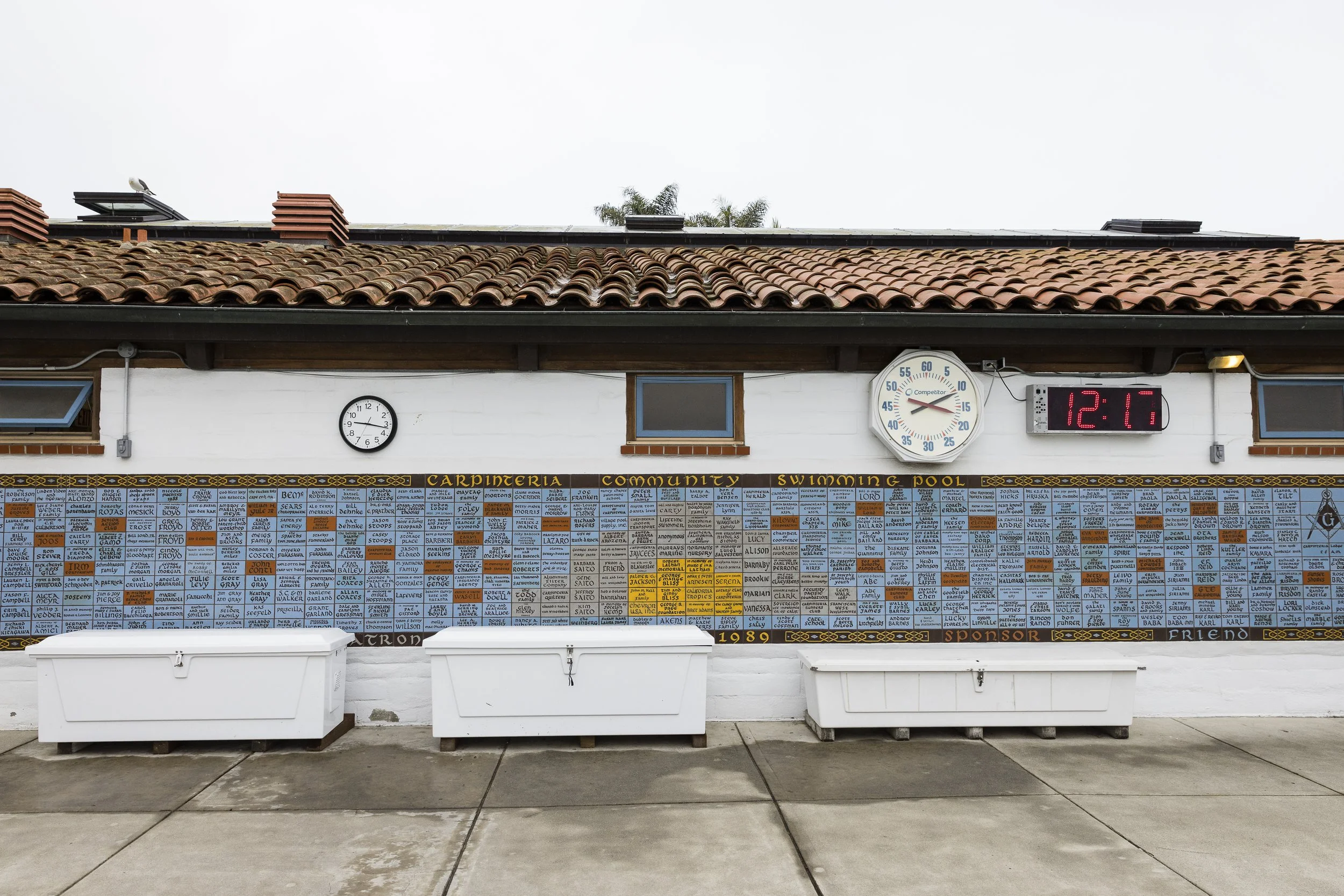 Wall with many colorful plaques with names, a small window, a clock, a large digital time display, and signs for carpentry and swimming pool.
