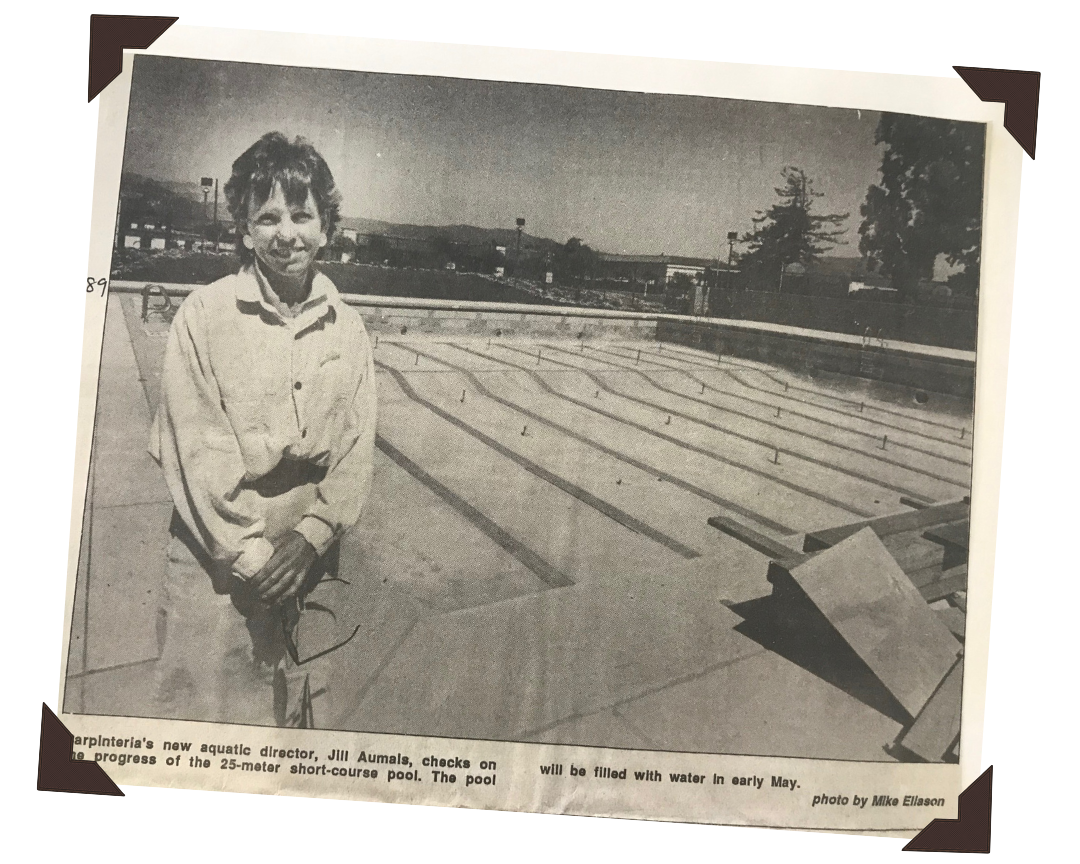 Black and white photo of a woman standing outdoors near a swimming pool with swimming lanes. She is wearing a light-colored jacket and holding sunglasses. There are trees and buildings in the background.