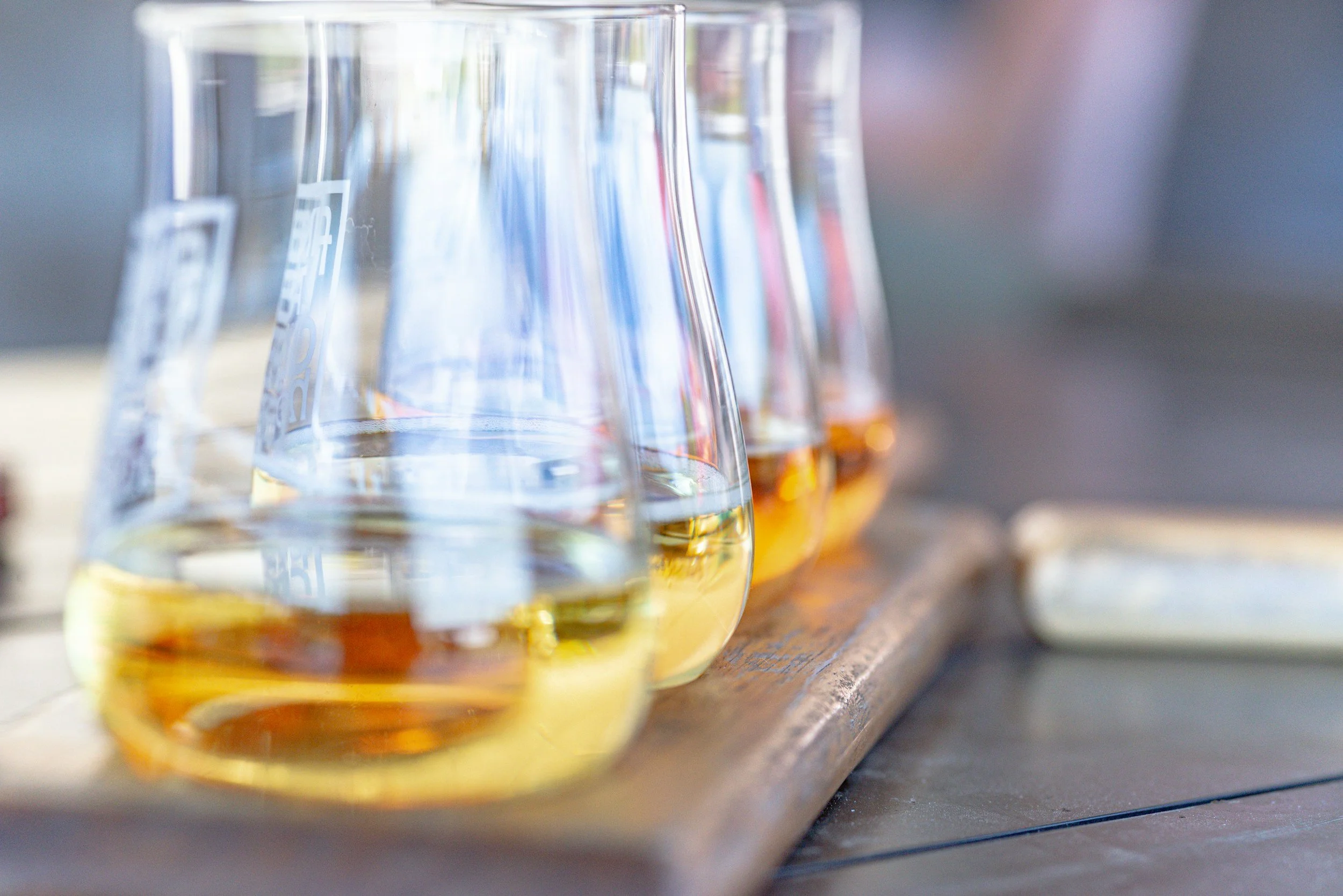 Close-up of empty whiskey glasses on a wooden surface, with some glasses partially filled with amber-colored liquid, in outdoor setting with blurred background.