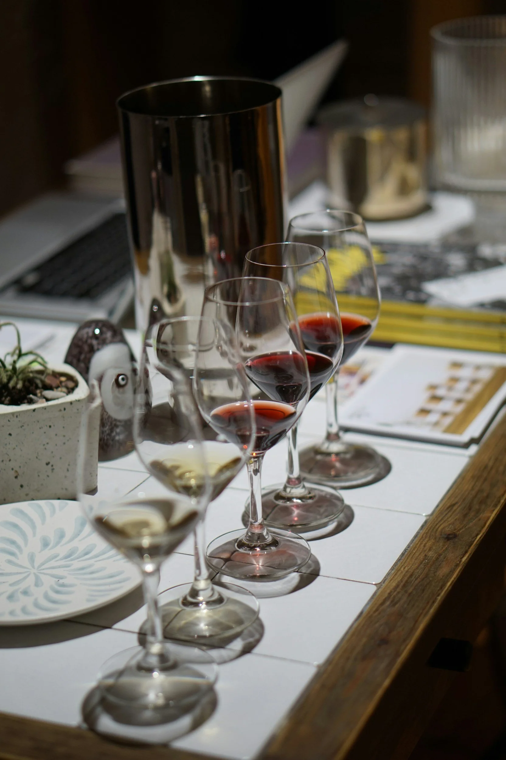A row of five wine glasses with red and white wine on a table, with a reflection in silverware and a blurred background including a napkin holder and a plant.