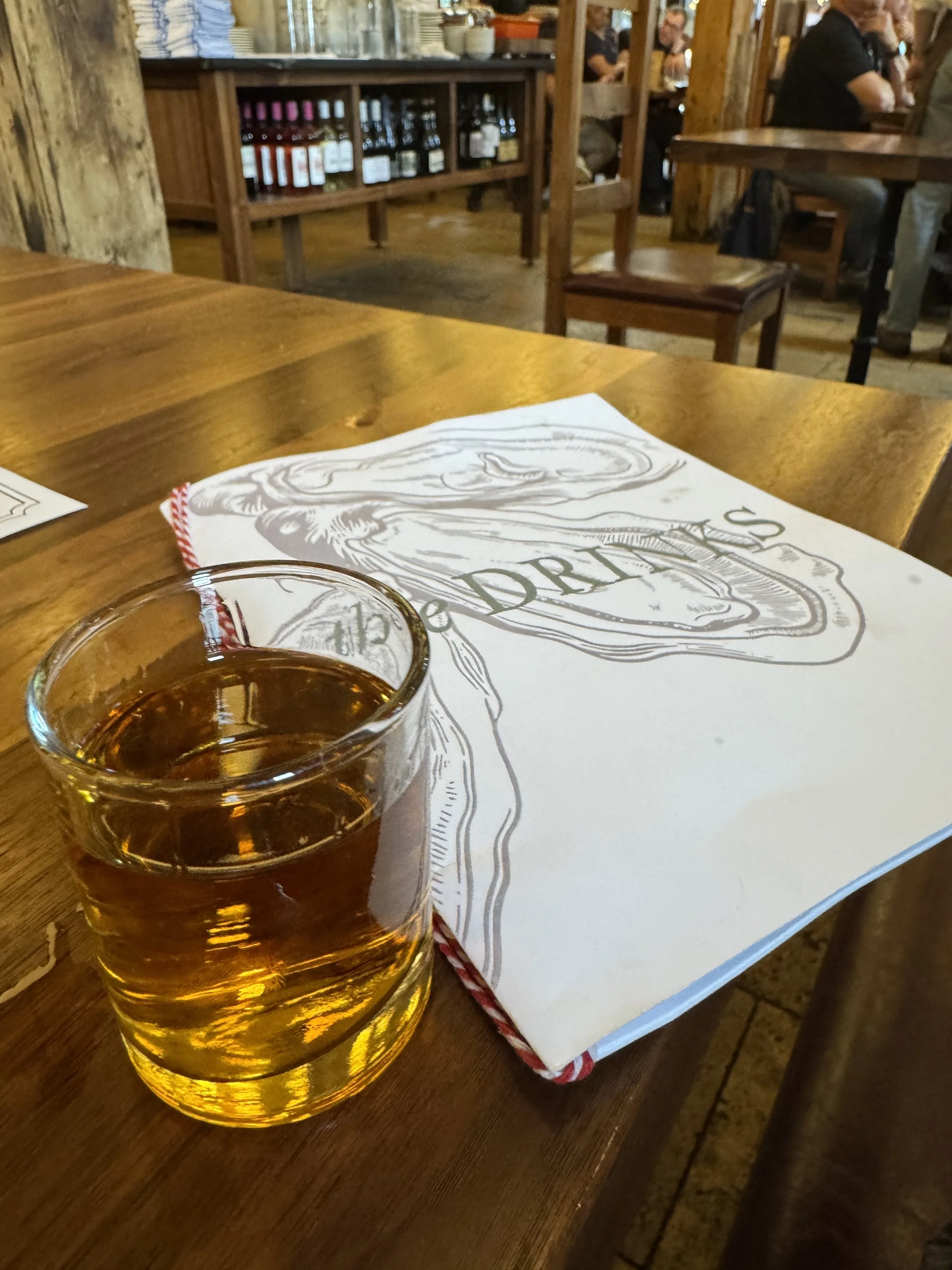 A glass of amber-colored drink on a wooden table in a rustic restaurant setting with chairs, a paper napkin, and a wooden shelf with bottles in the background.