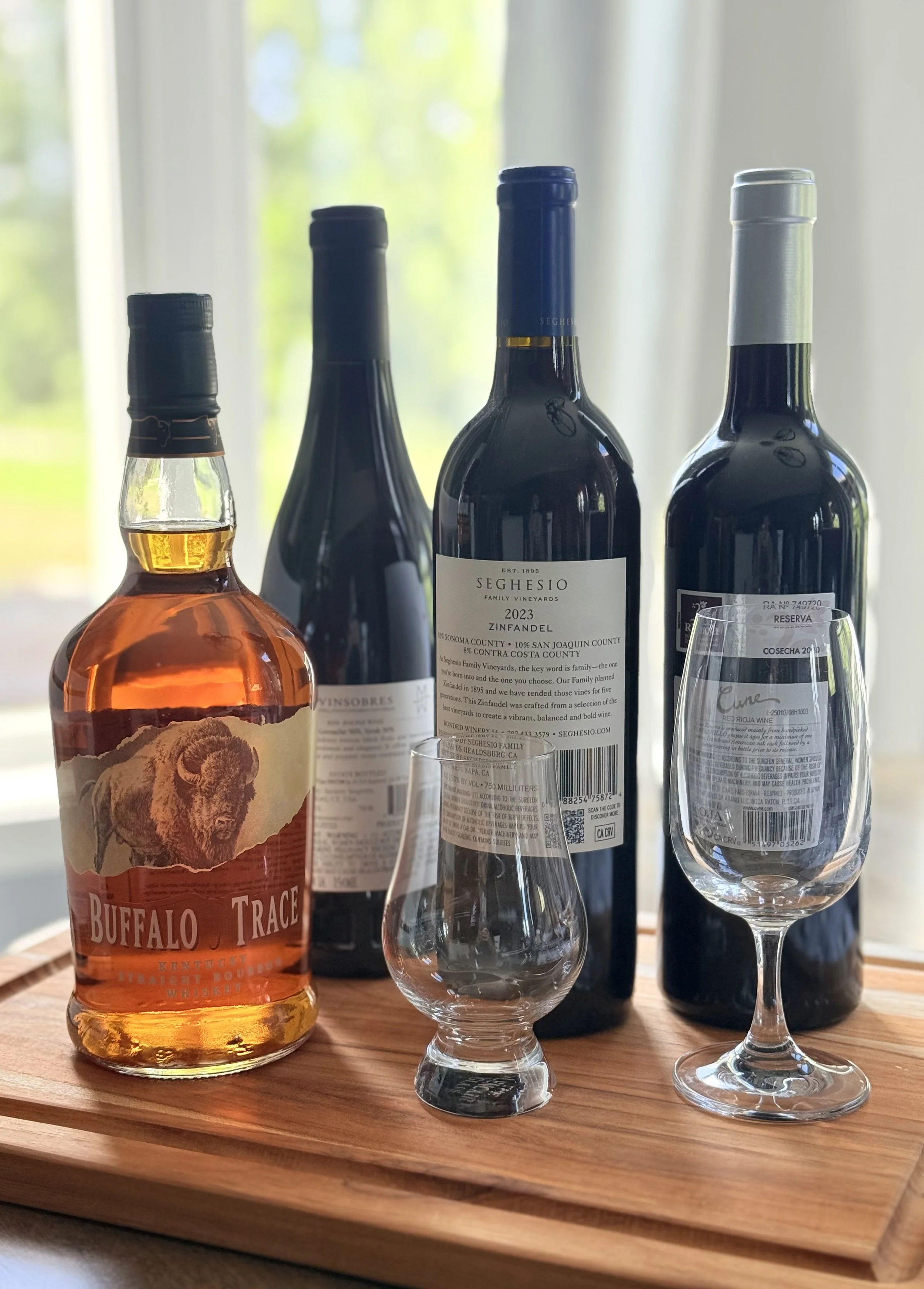 Four bottles of wine and a glass of red wine on a wooden tray with a window in the background.