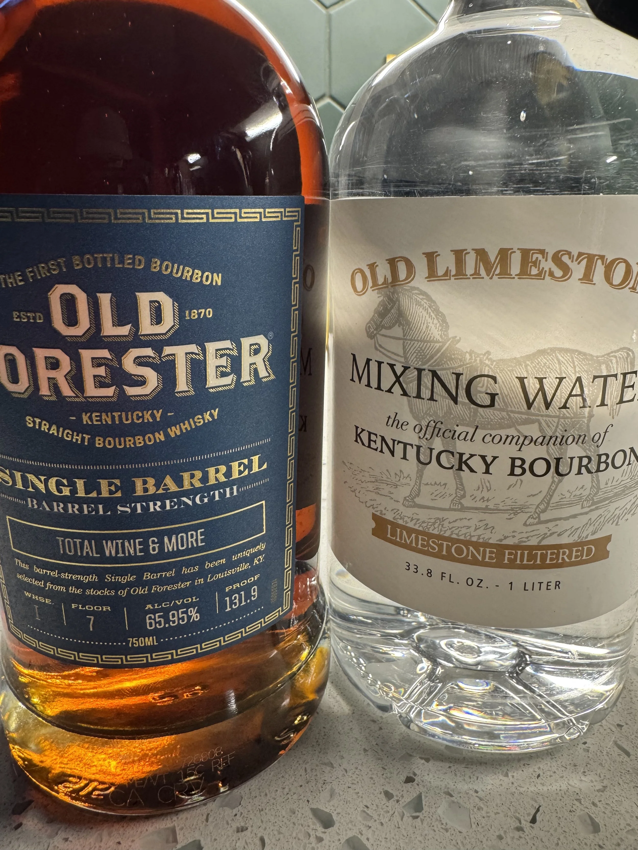Close-up of a bottle of Old Forester bourbon whiskey and a bottle of Old Limestone Filtered Water on a grey speckled countertop.