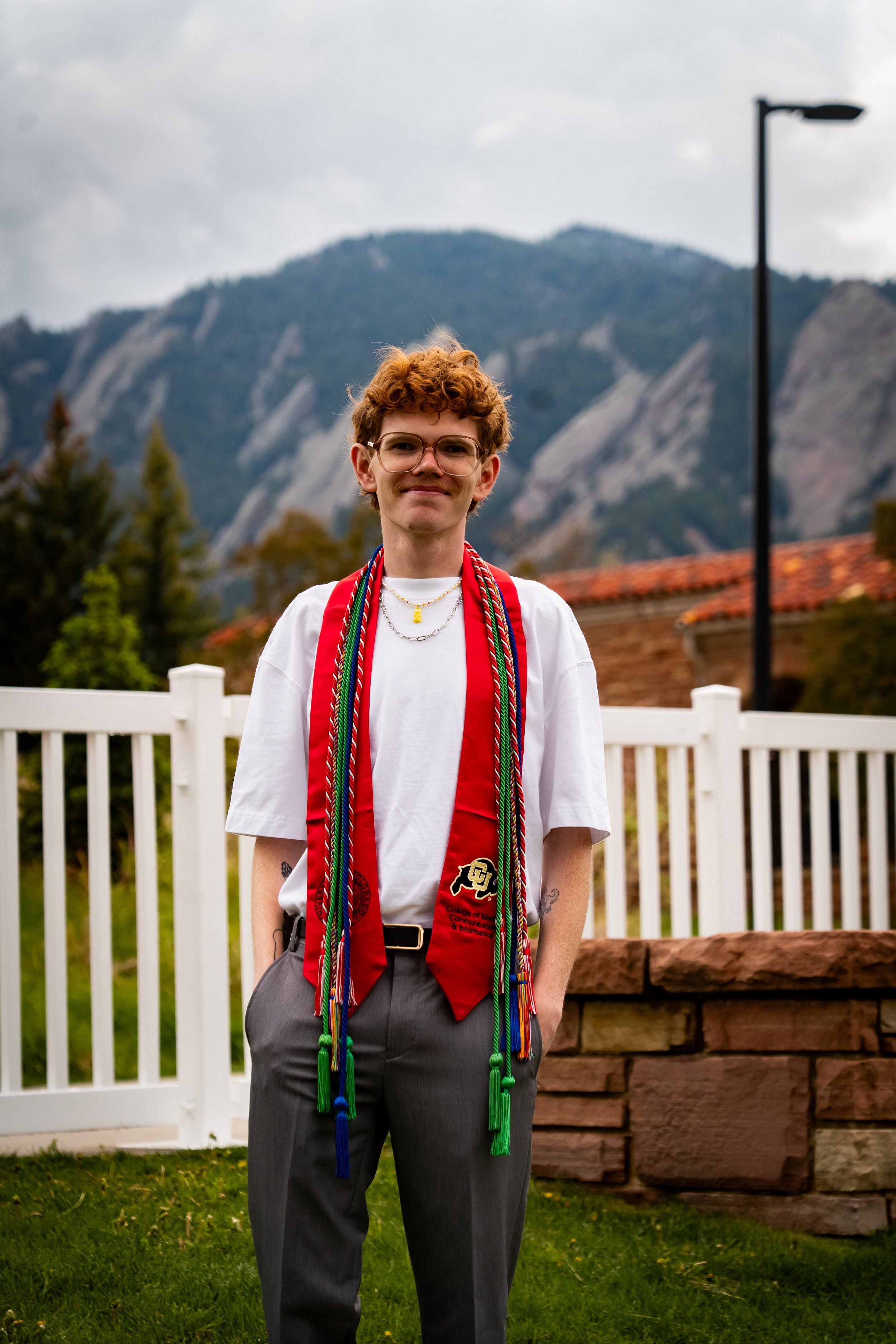 Young man with curly red hair and glasses standing outdoors with a mountain in the background, wearing graduation cords and an honor stole with university logos, in front of a white fence and brick wall.