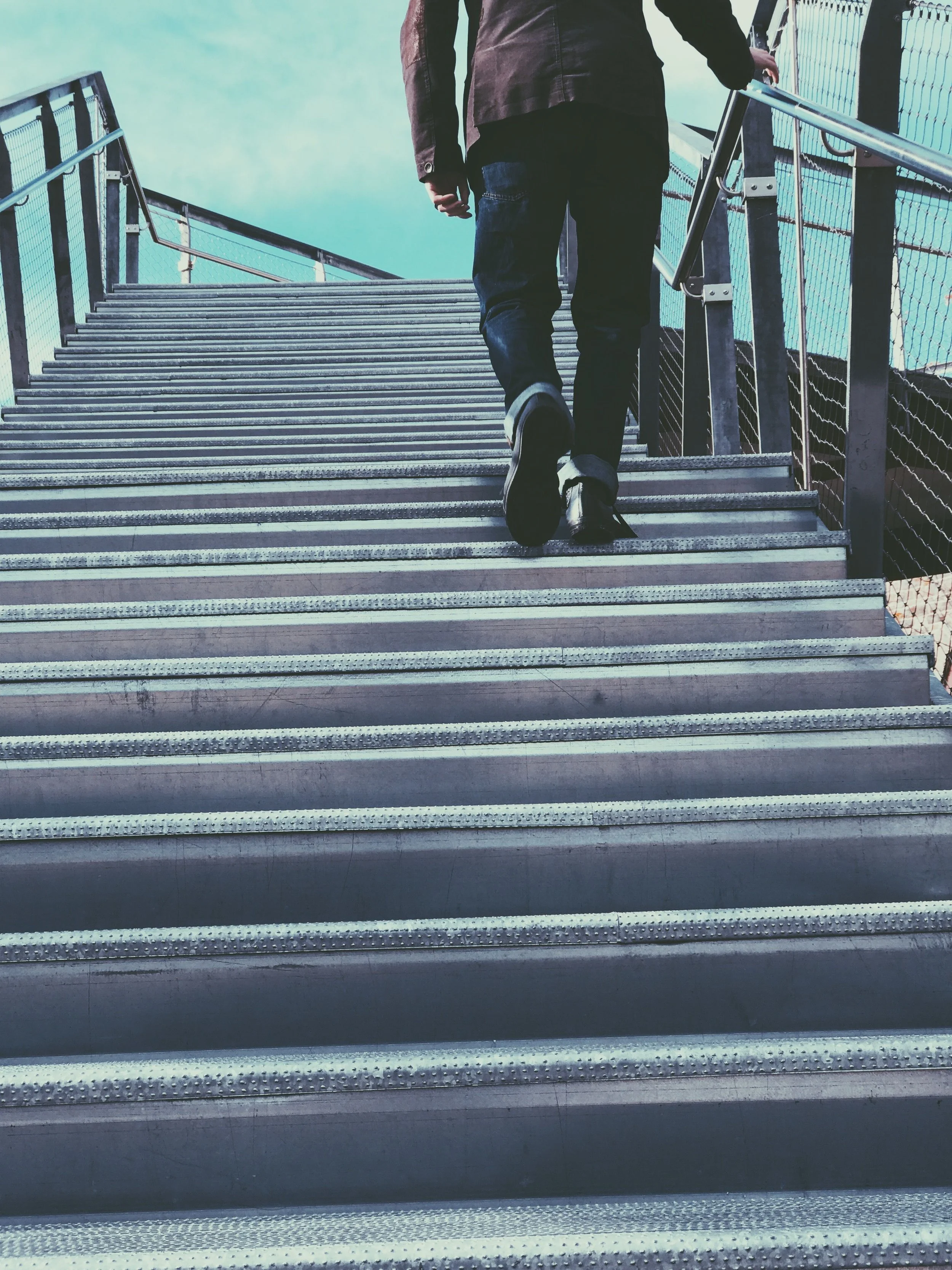 Person walking up outdoor metal stairs with railing, wearing black jeans and dark jacket.
