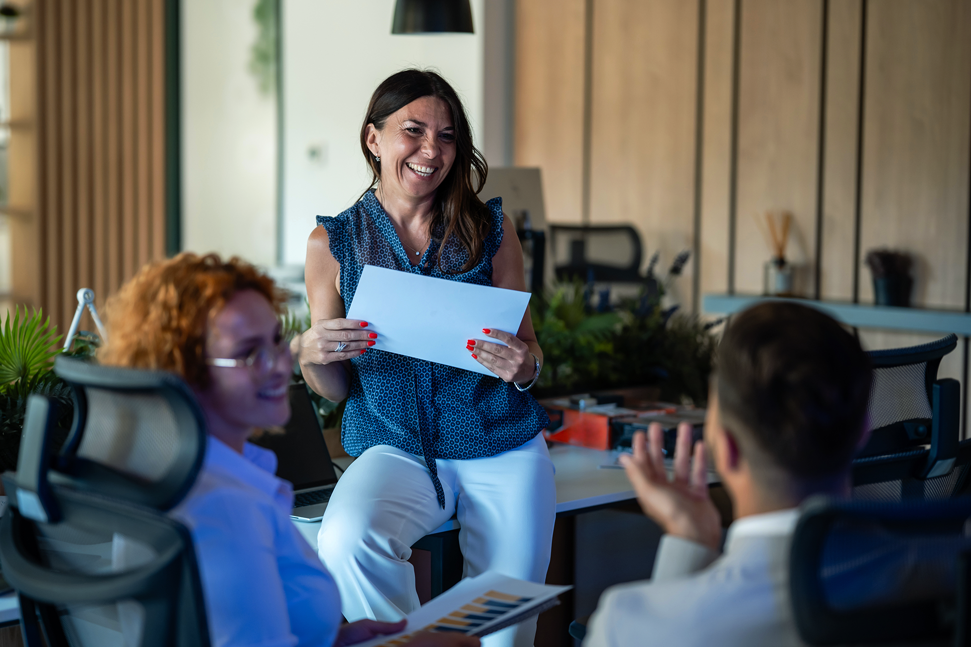 A woman with dark hair, wearing a blue patterned sleeveless top, is standing and smiling, holding a piece of paper as she interacts with seated individuals in a modern office setting.
