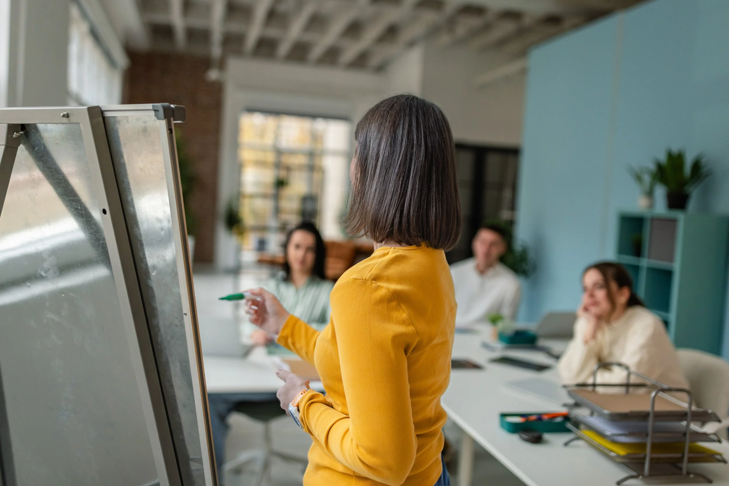 A woman with shoulder-length brown hair wearing a yellow long-sleeve shirt standing in front of a whiteboard and writing with a green marker in a modern office or conference room. Other people are visible in the background, sitting and listening.