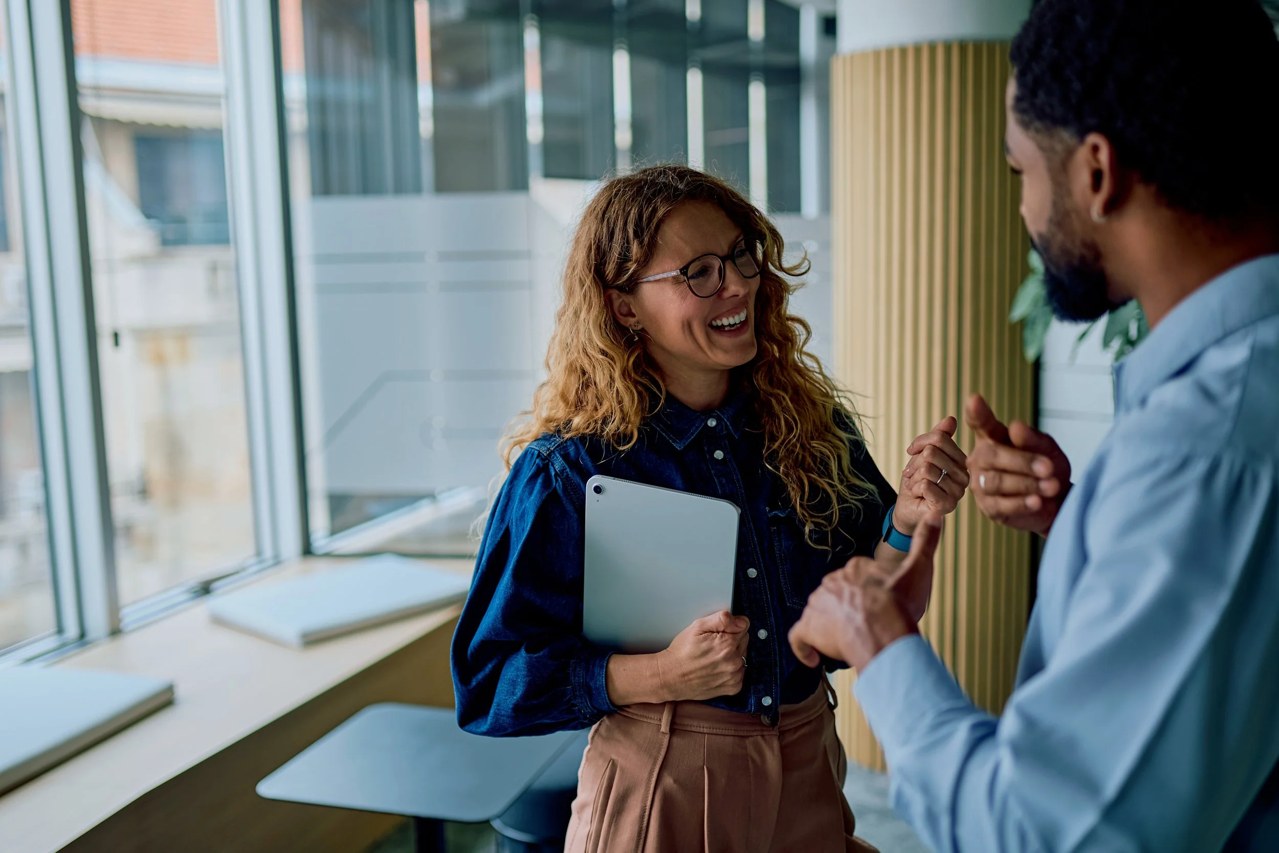 A woman with curly red hair, glasses, and a denim shirt smiling and holding a tablet, shaking hands with a man with dark hair and a beard, wearing a blue shirt, in a modern office setting with large windows and green plants.