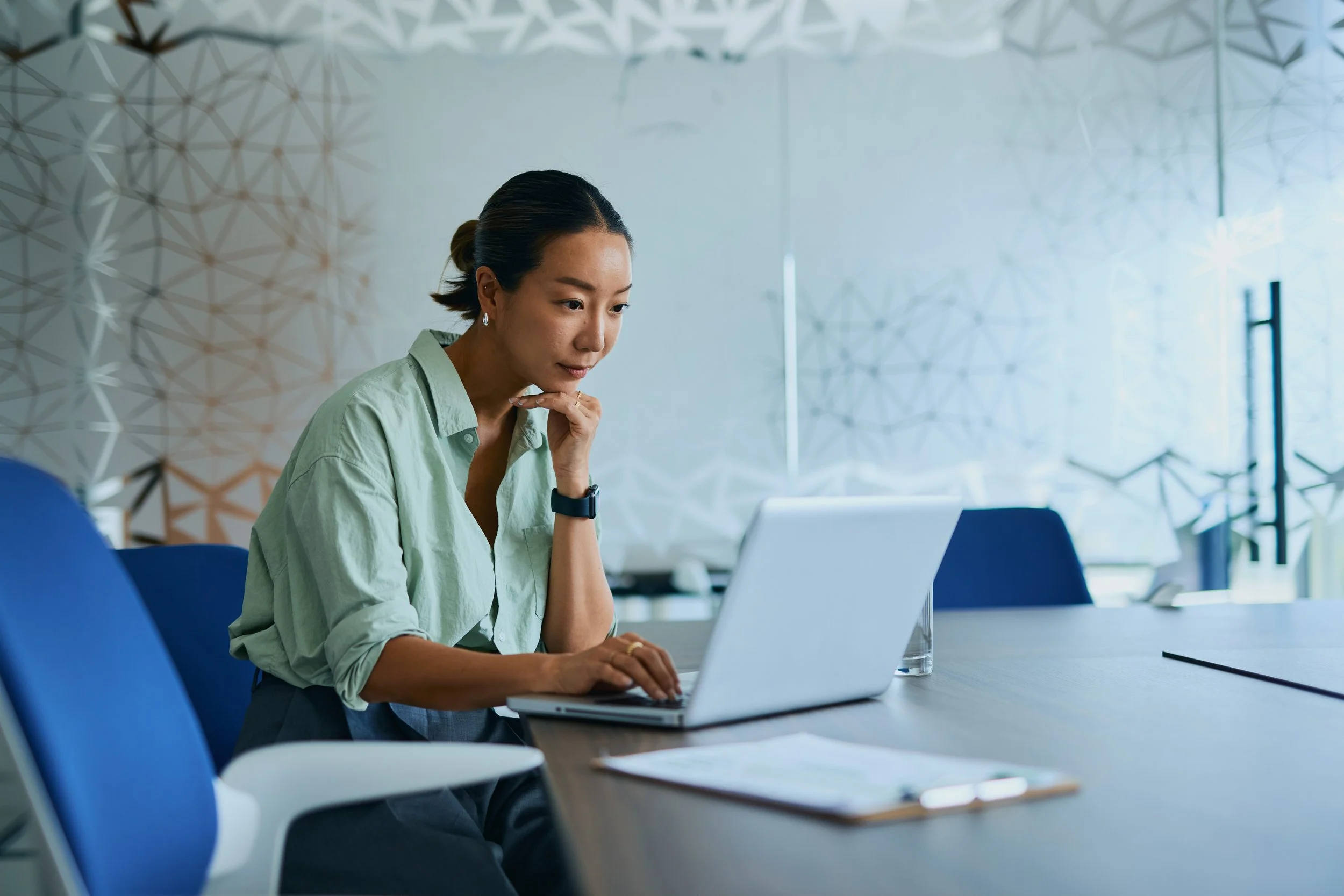 A woman with dark hair tied back, wearing a light green shirt, sitting at a desk working on a laptop in a modern office space. She is looking at her laptop screen with her right hand near her chin, appearing focused. The background features a geometric patterned wall and bright natural light.