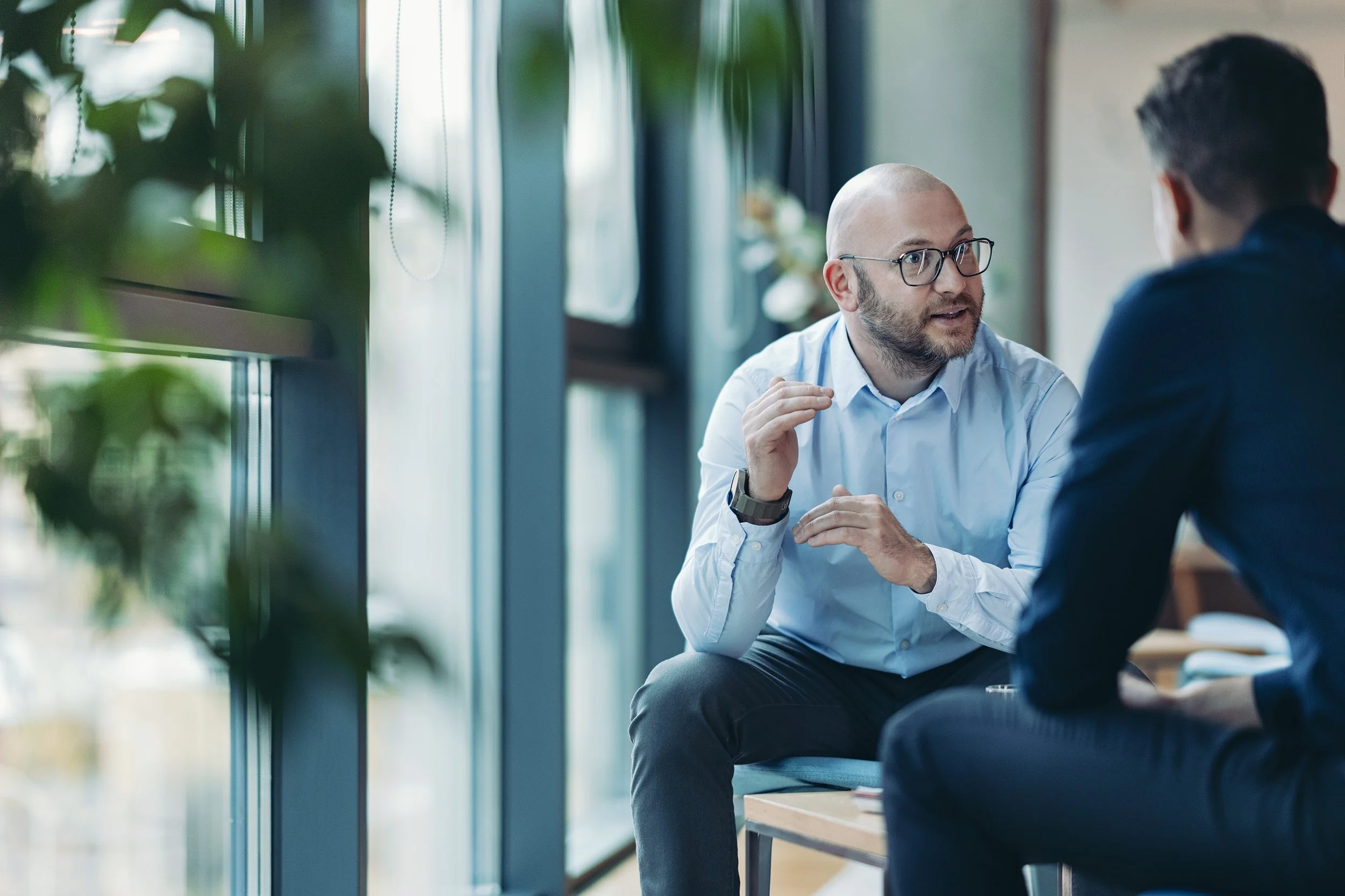 Two men are having a conversation in a modern office setting, one is bald with glasses and a light blue shirt, the other has dark hair and is wearing a dark jacket. The man in glasses is speaking and gesturing with his hand.