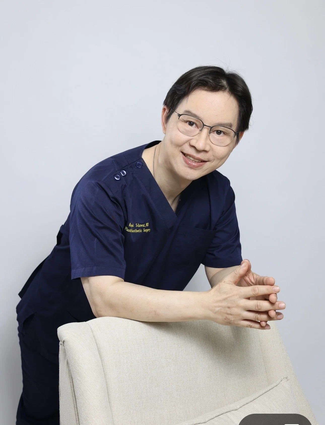A male doctor in a navy blue medical uniform with glasses smiling and leaning on a beige chair.