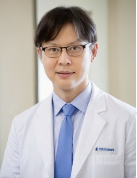A male doctor wearing glasses in a white lab coat and blue tie, standing indoors with a neutral background.