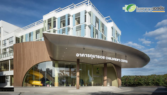 Modern children's center building with glass windows and a curved wooden facade, with the sign in Thai and English reading 'Children's Center' and a hospital logo in the top right corner.