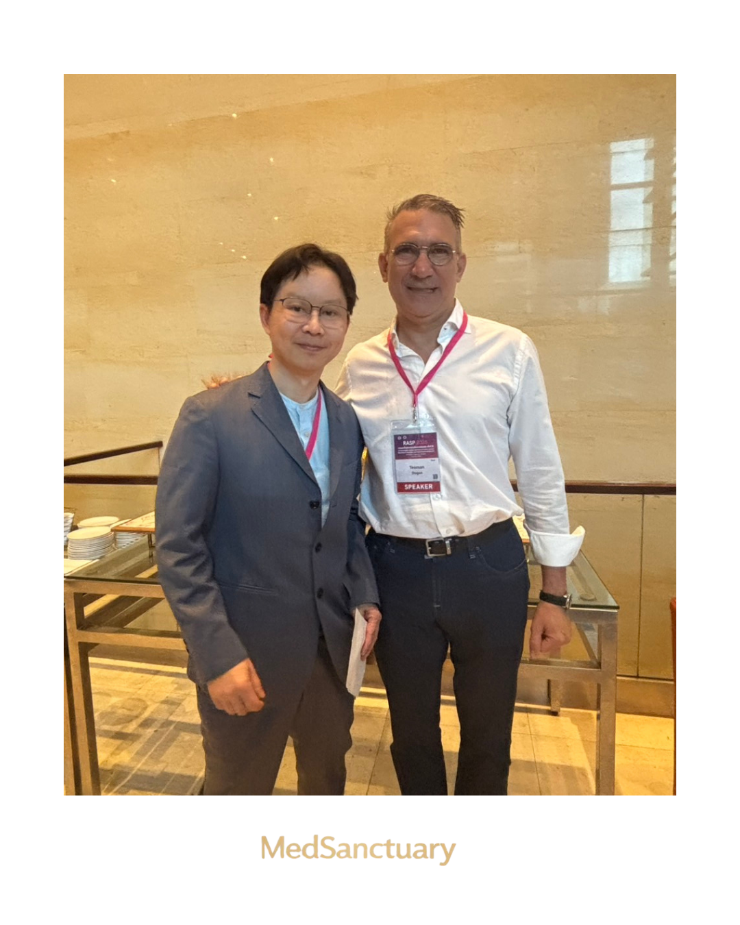 Two conference attendees pose together indoors, with one wearing a speaker badge and the MedSanctuary logo below.