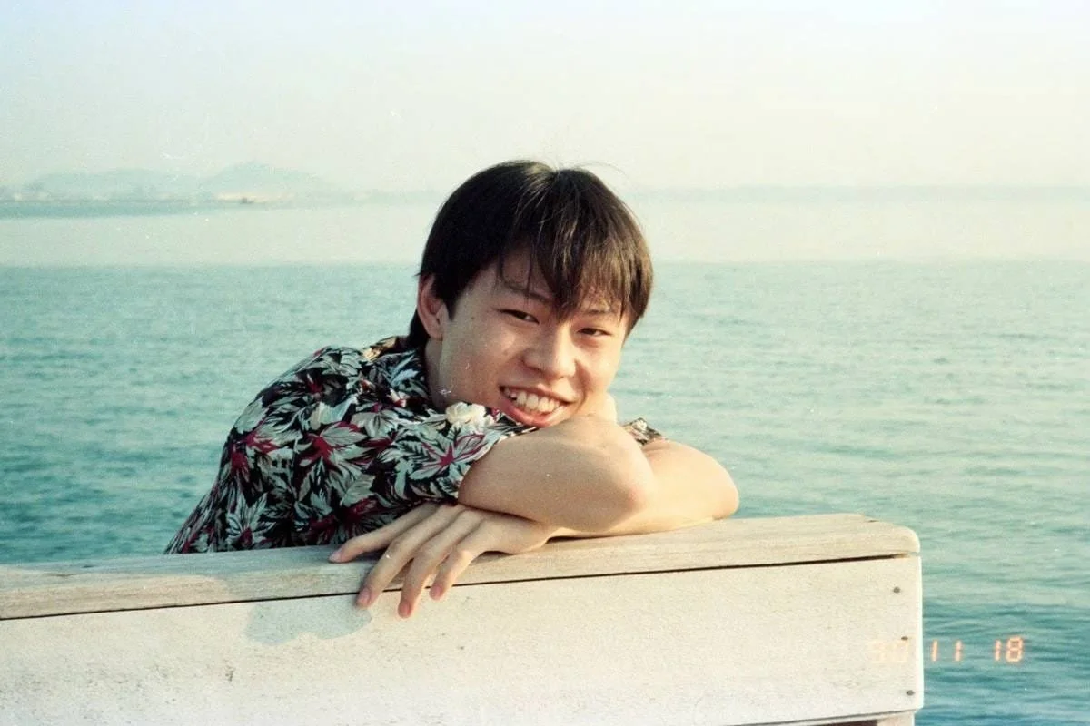 Young man in a floral shirt leaning on a boat railing with the ocean in the background