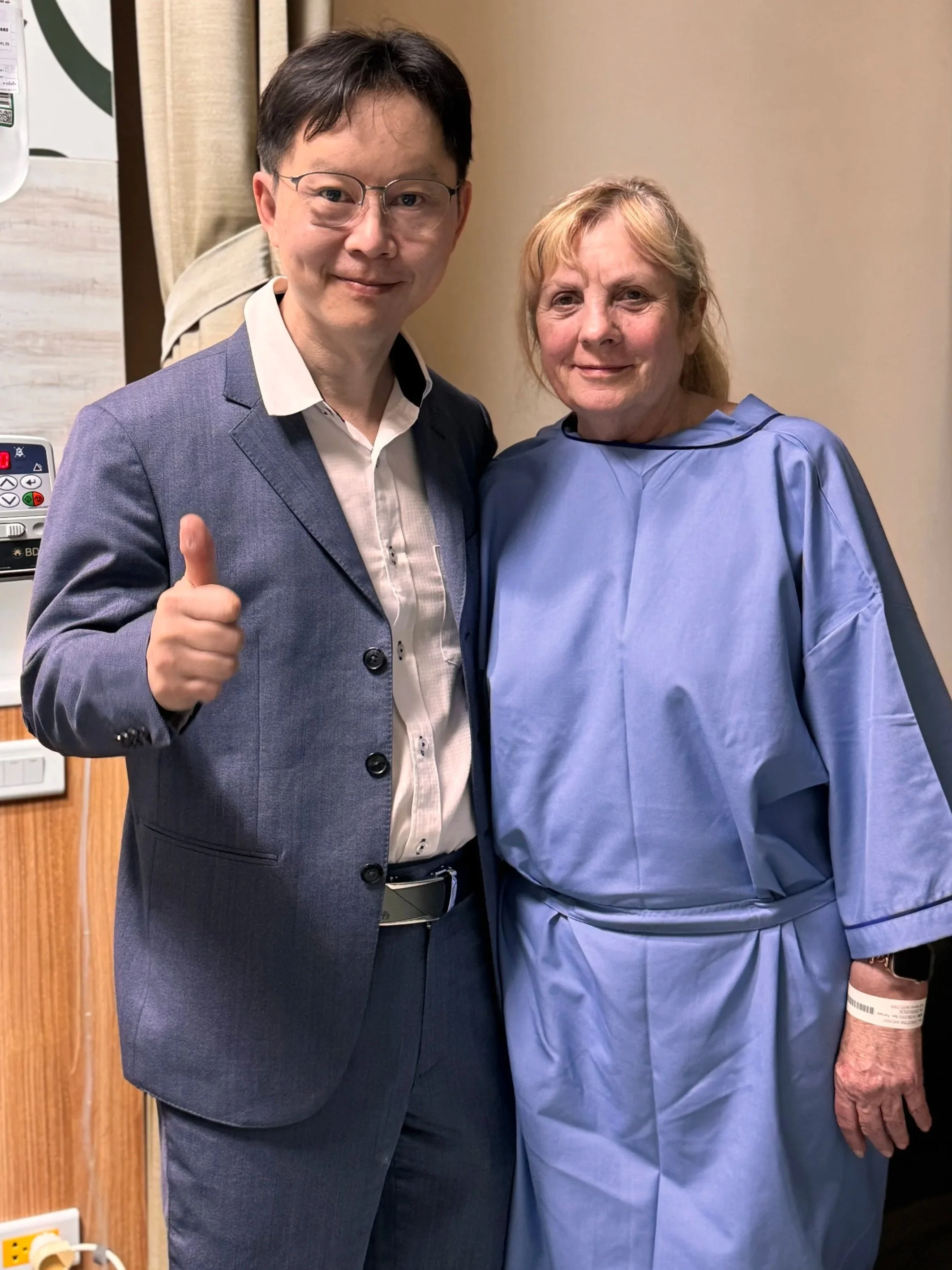 Man in a suit giving a thumbs-up beside a smiling woman in a hospital gown