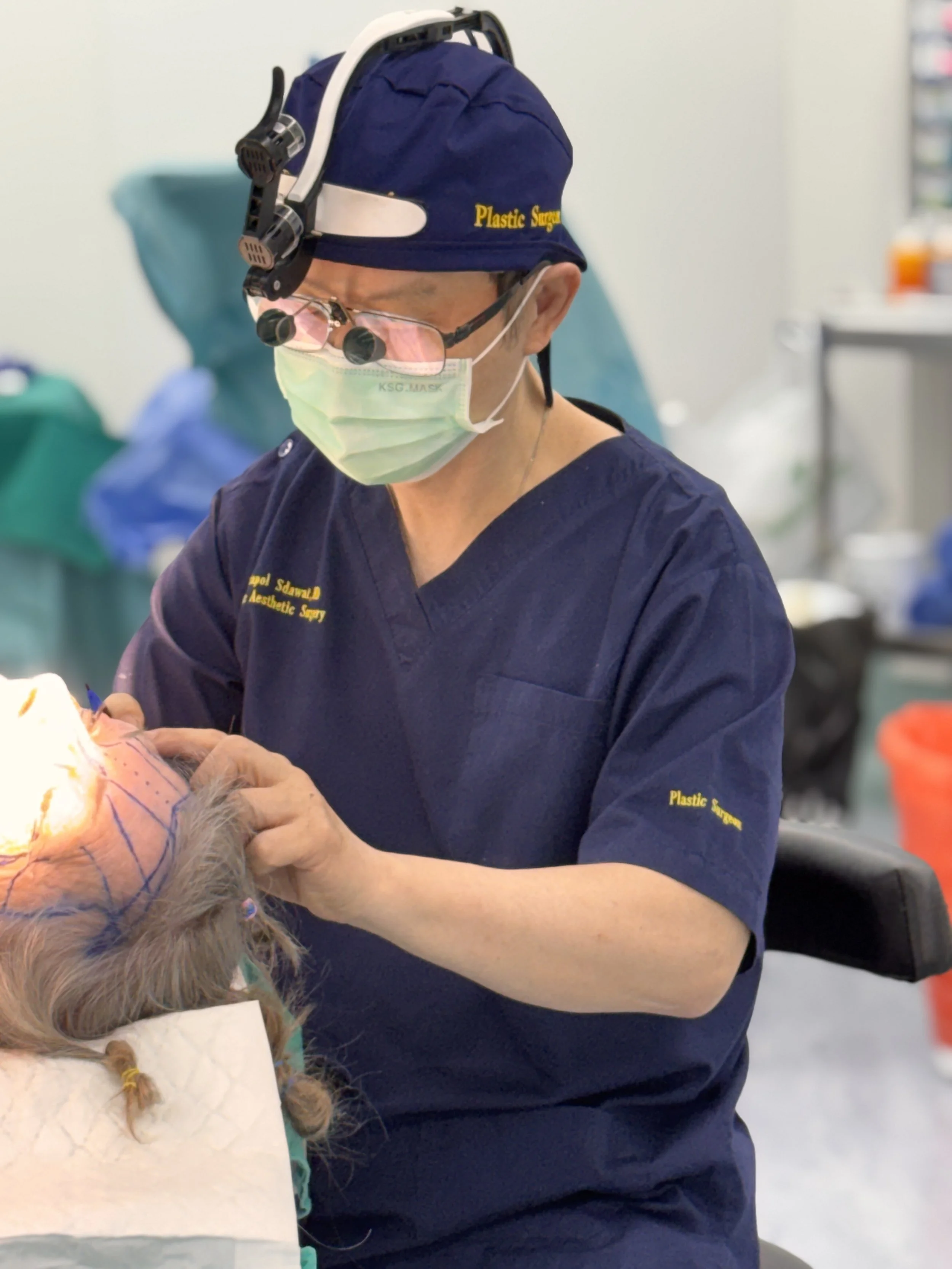 Plastic surgeon in scrubs, surgical mask, magnifying loupes, and headlamp performing a procedure on a patient