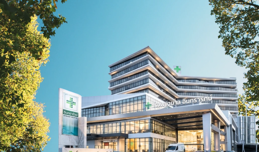 Modern multi-story hospital building with glass windows and green medical cross logo, surrounded by trees during daytime.