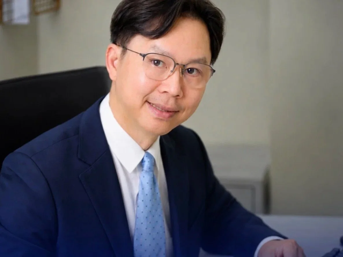 A man wearing glasses, a dark blue suit, white shirt, and light blue tie, sitting at a desk in an office, smiling at the camera.