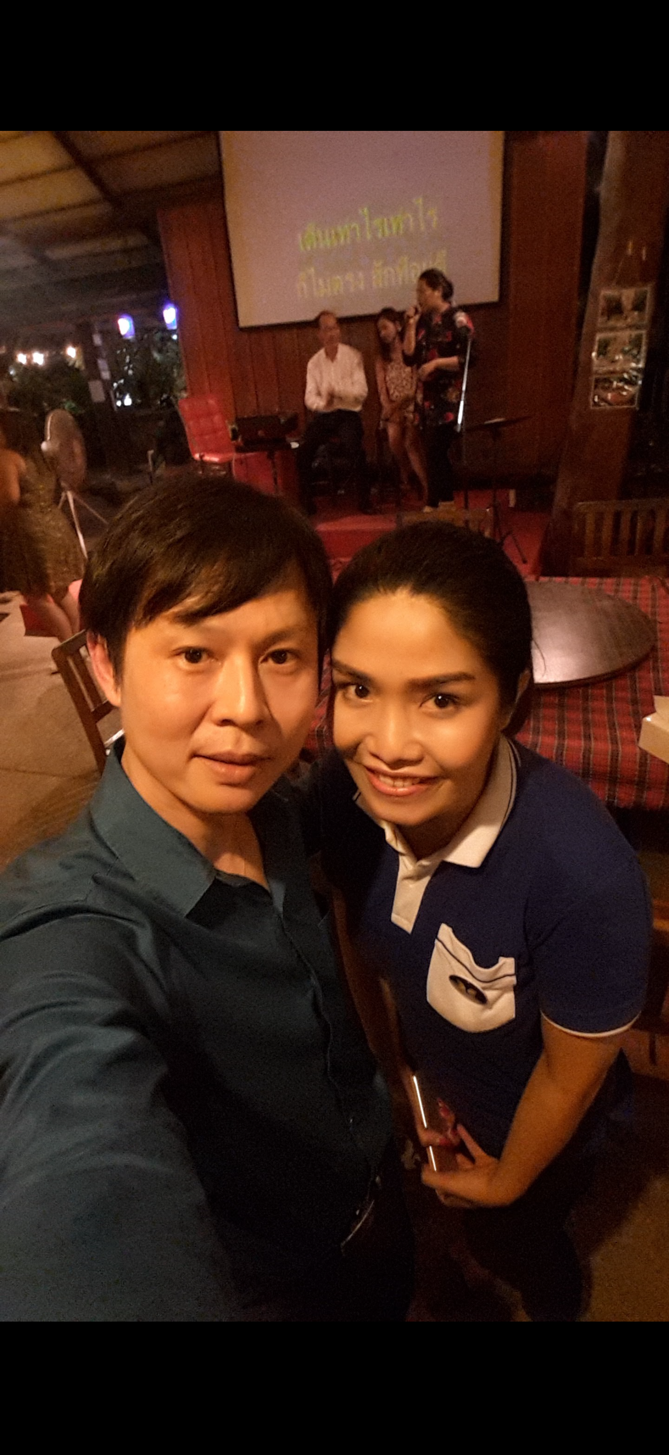 Two people take a selfie at a restaurant or event while performers stand onstage behind them.