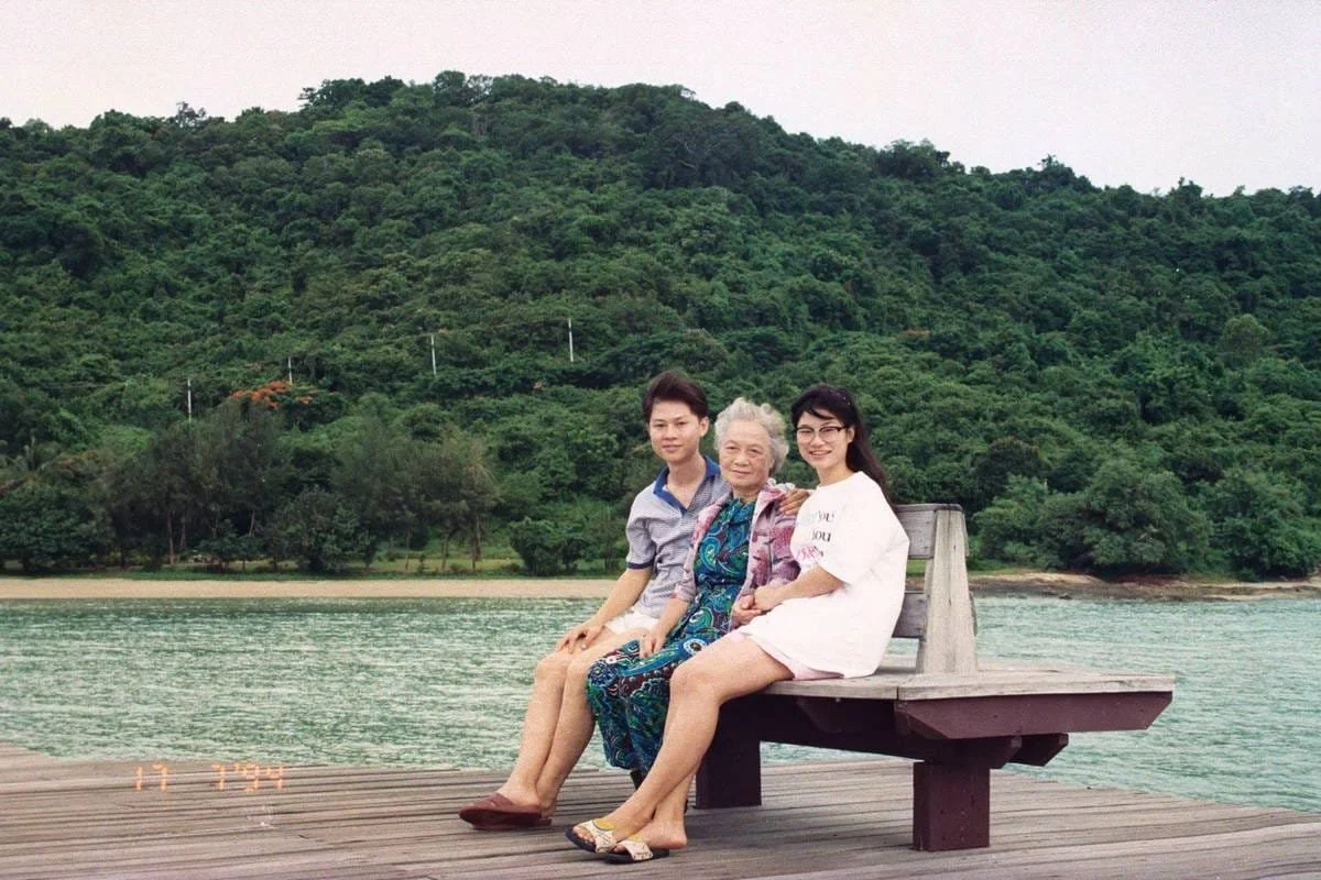 Three women sitting on a bench beside turquoise water, with a forested hillside in the background.