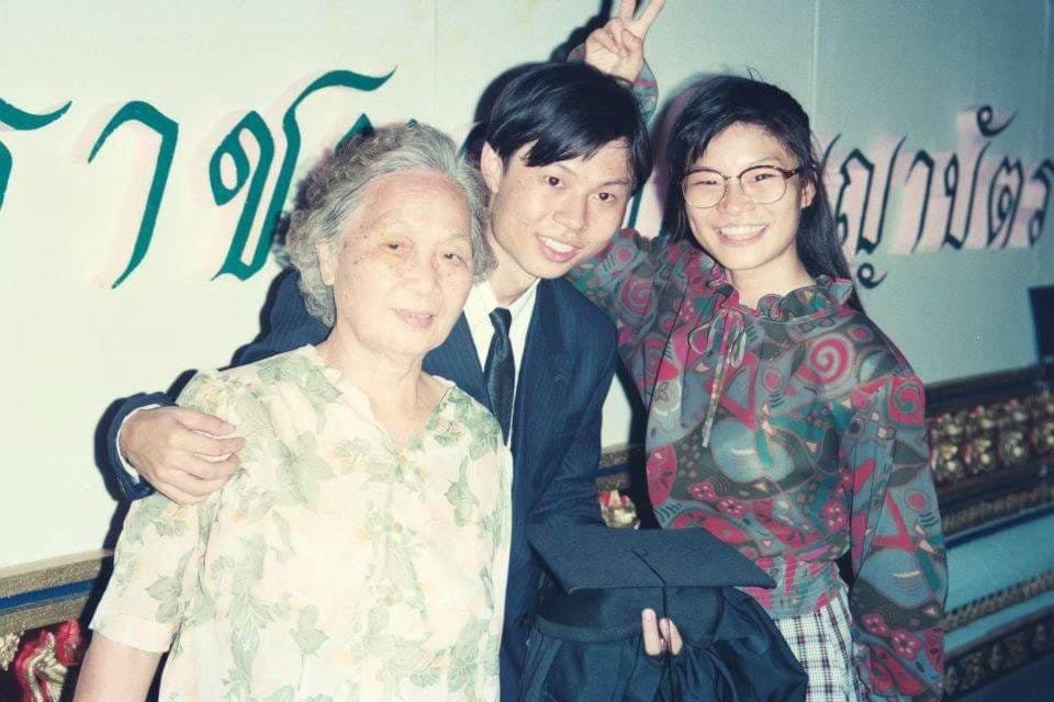 Three people posing together indoors, with a person making bunny ears behind them and decorative script on the wall.