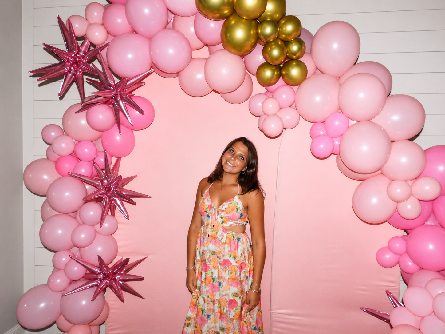 A woman in a colorful floral dress standing in front of a pink balloon backdrop with star-shaped balloons and various sized pink and gold round balloons.