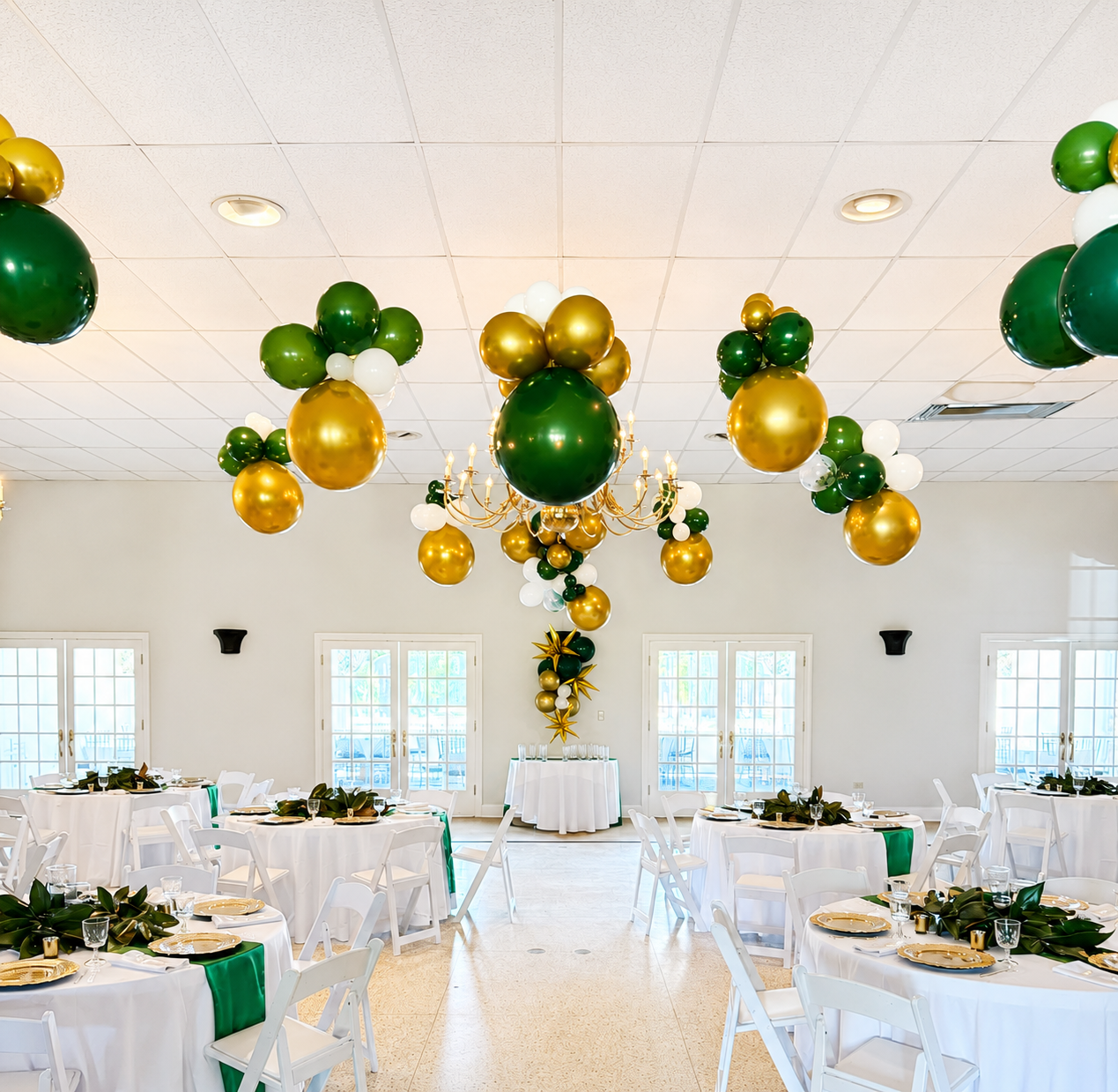 Decorated banquet hall with white tables and chairs, featuring green, gold, and white balloons hanging from the ceiling, with a central balloon arrangement and crystal chandelier.