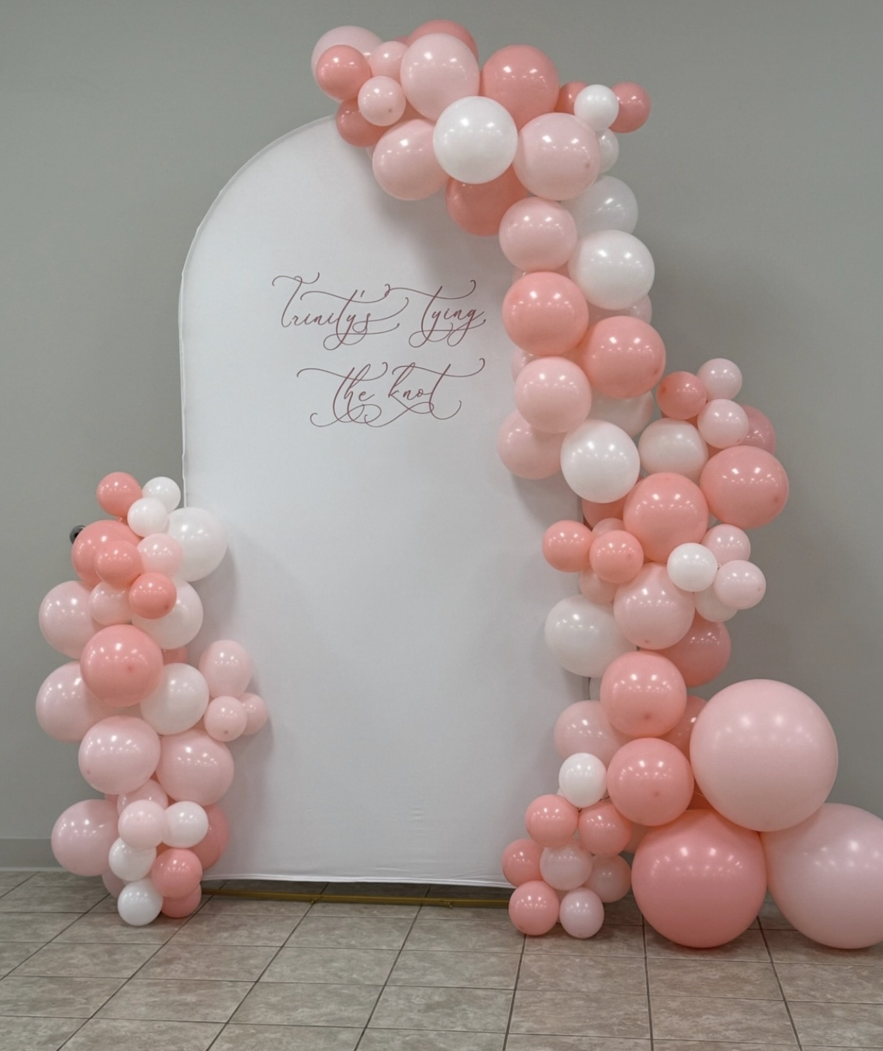 Decorative backdrop with pink and white balloons arranged around a white board with the text 'Trinitys Tugs the Knot' written on it.