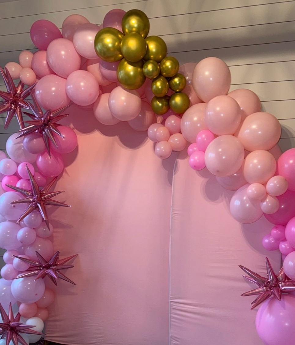 A balloon arch decorated with pink, white, gold, and rose gold balloons and star-shaped balloons, arranged against a pink backdrop.