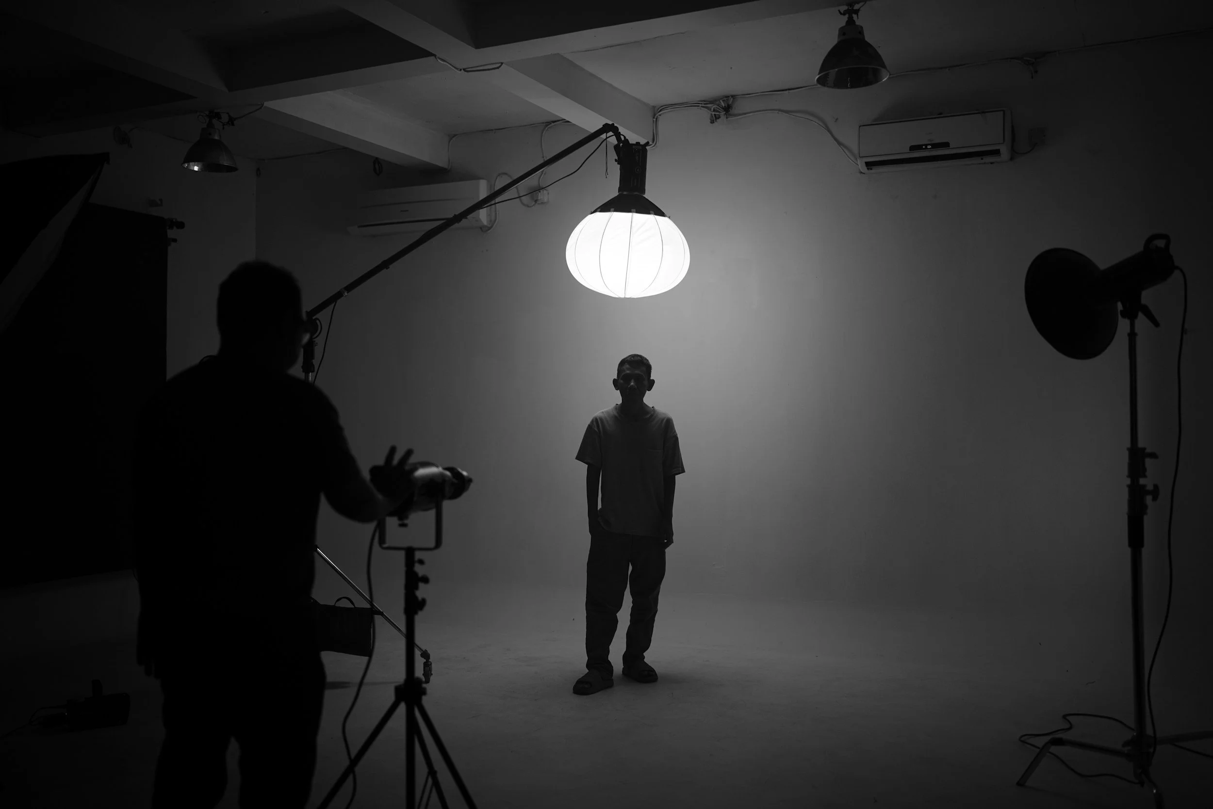 A black-and-white photo of a photo studio setup with a young boy standing in front of a plain background, lit by a large overhead light. Several crew members and studio equipment are visible preparing for a photo shoot.