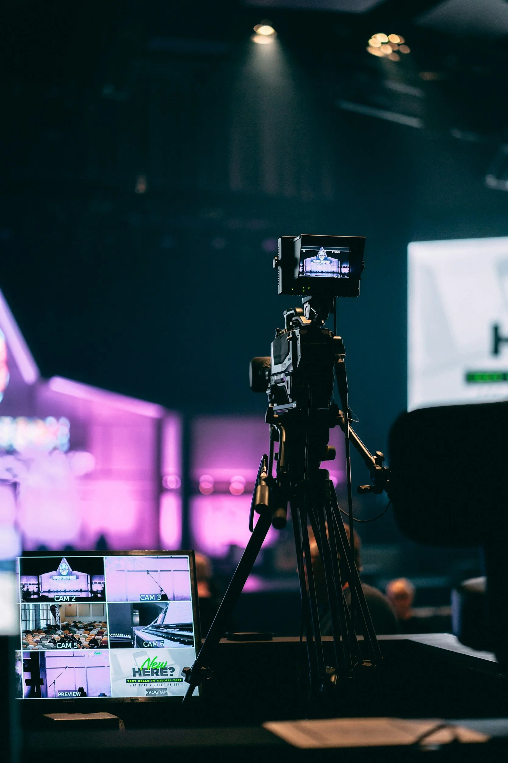 A professional video camera on a tripod captures footage at an indoor event with purple and white lighting. A monitor displays multiple camera feeds and graphics.