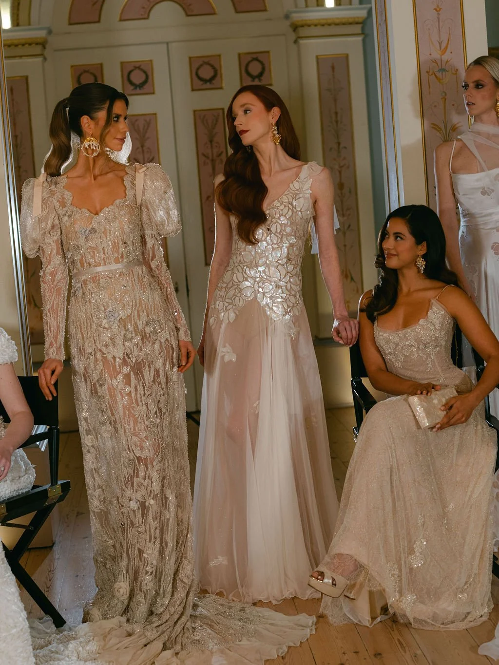 Four women in ornate dresses, possibly at a fashion show or bridal event, inside a decorated room with historical design elements.