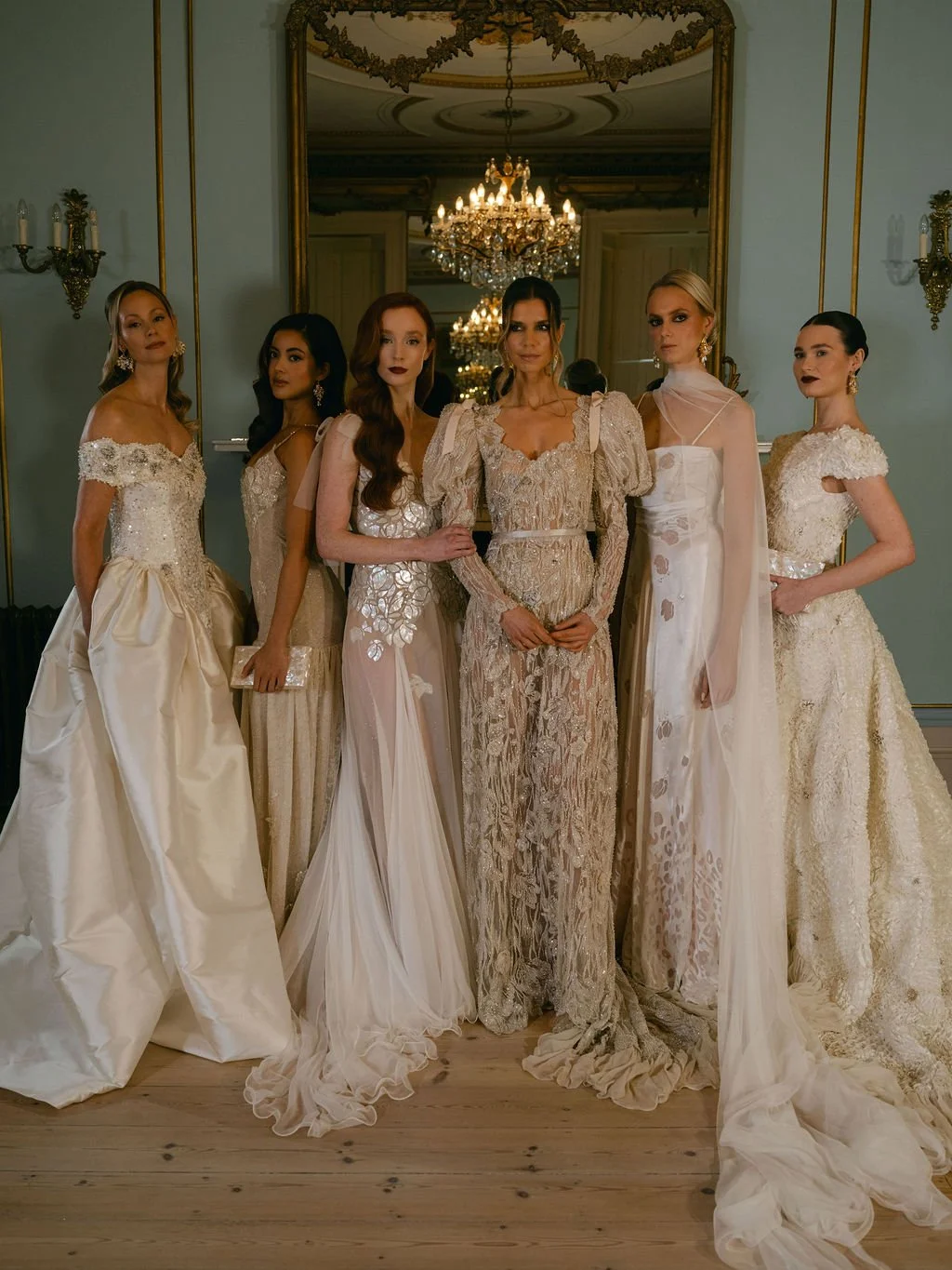 Group of six women in elegant, bridal-style dresses posing in a luxurious room with chandelier, large mirror, and ornate wall sconces.