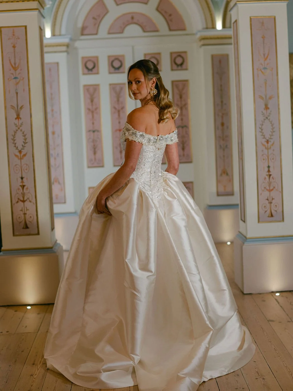 A woman in a wedding dress standing in an elegantly decorated room with pastel-colored walls and ornate paneling.