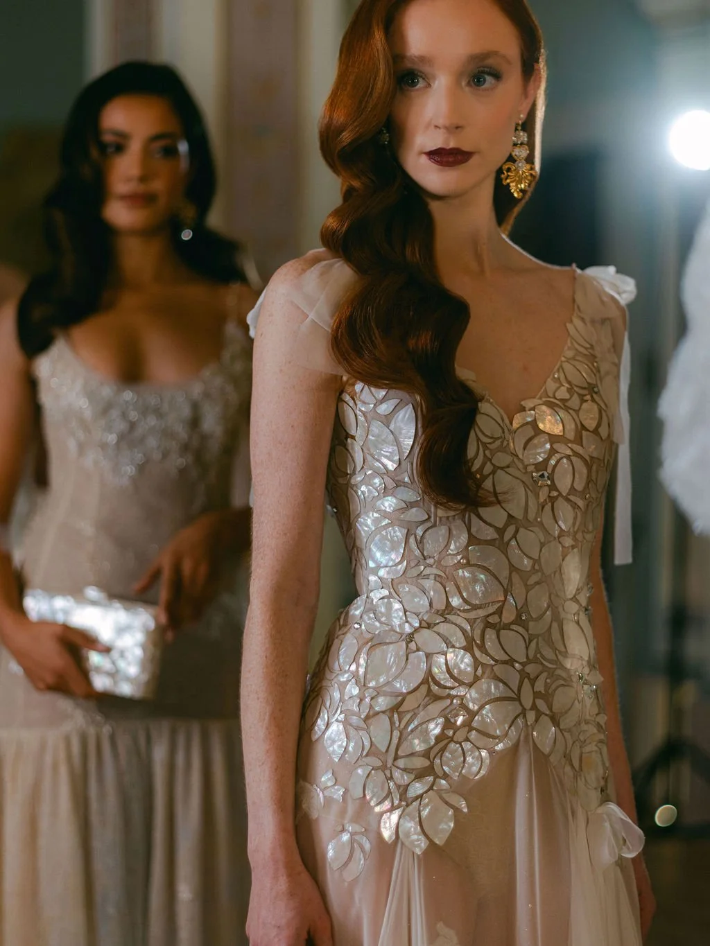 Women in elegant dresses at a formal event, one woman with long, wavy red hair and dark lipstick in the foreground, another woman in the background with dark hair and a beaded gown.